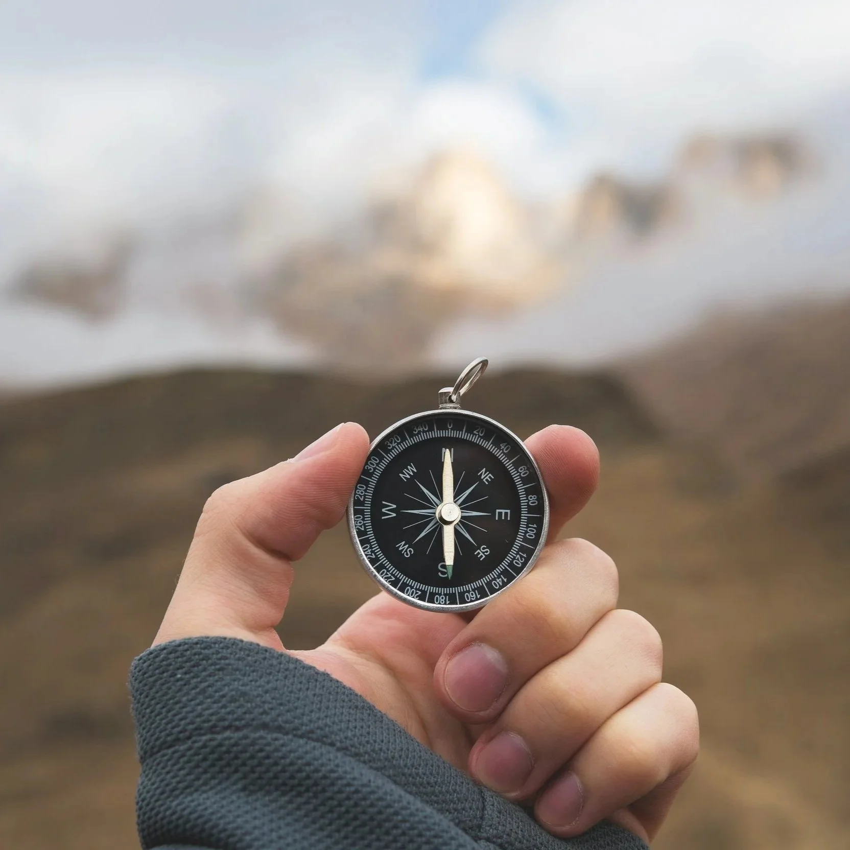 Hand holding a compass outdoors with a blurred mountain landscape in the background.