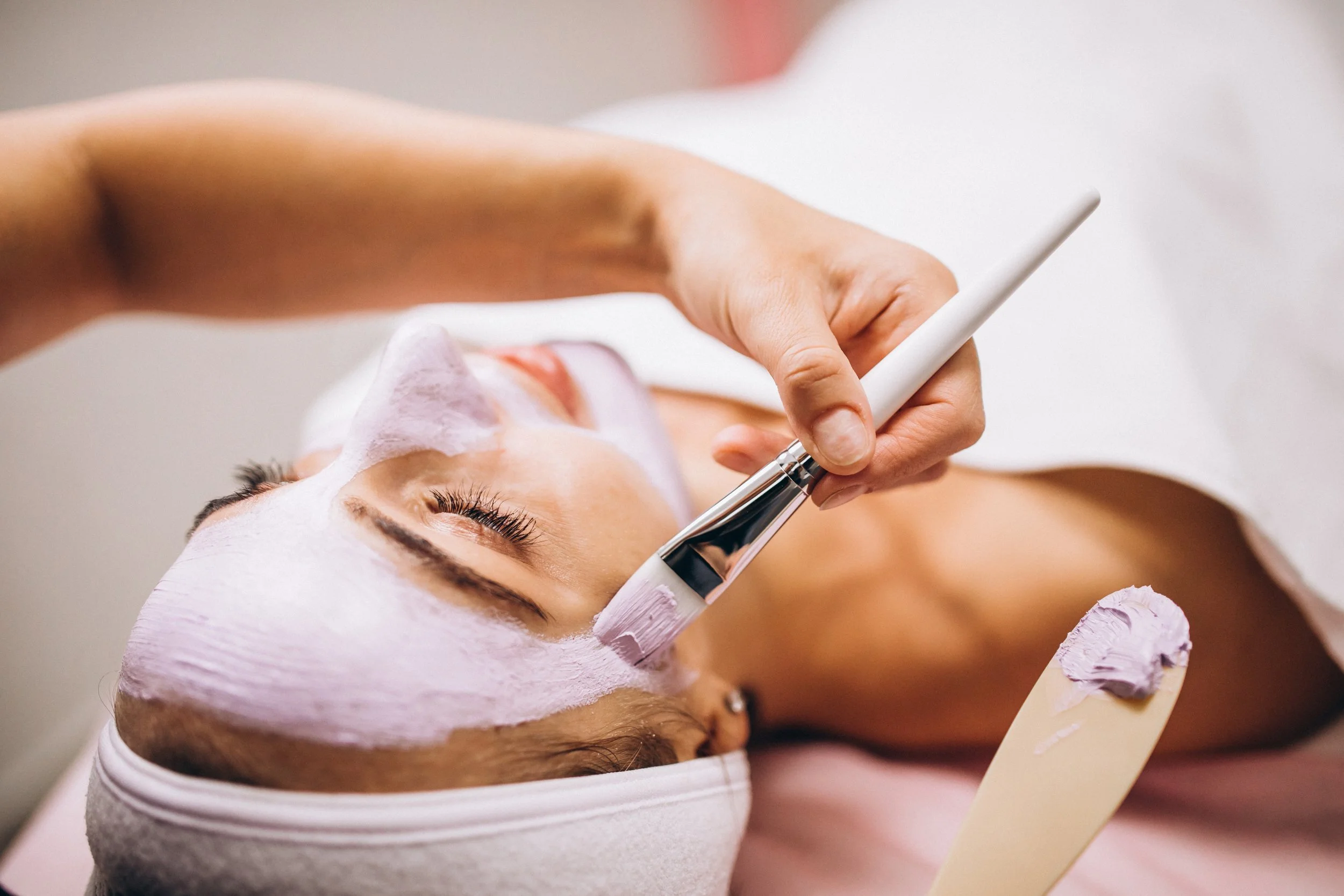 Una mujer recibiendo una limpieza facial en un spa, con una mascarilla en la cara y una brocha aplicada por una esteticista.
