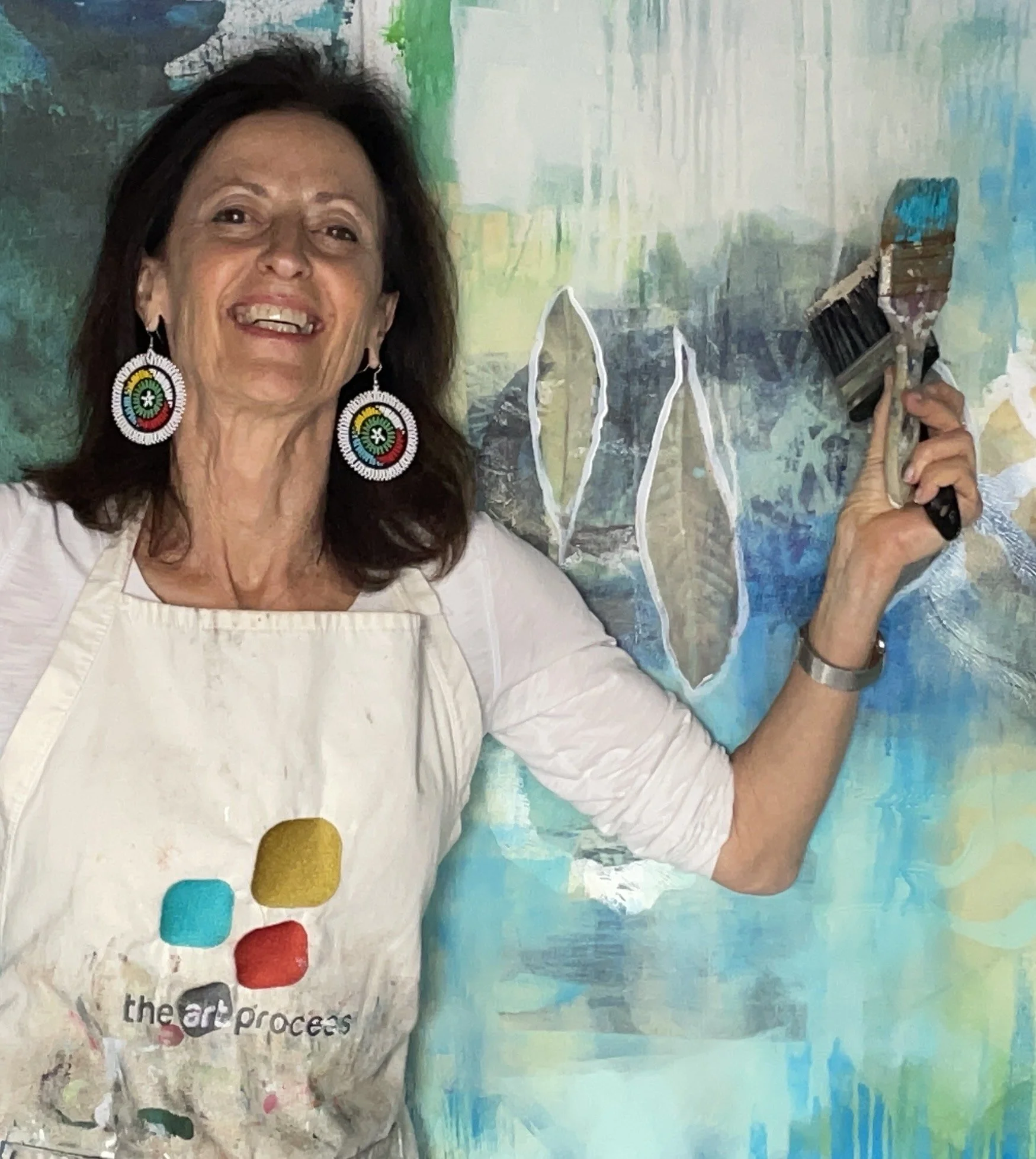 A woman smiling while holding a paintbrush in front of a colorful abstract painting. She is wearing large, colorful earrings and a white apron with the words 'the art process' on it.