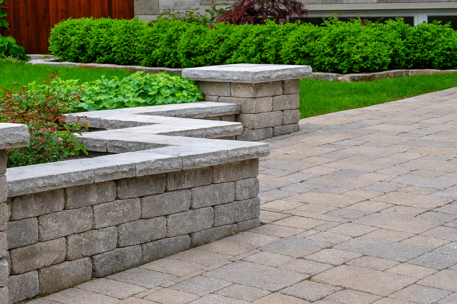 Stone garden wall with flat top stones, surrounding a flower bed with green plants and shrubs, adjacent to a paved walkway.