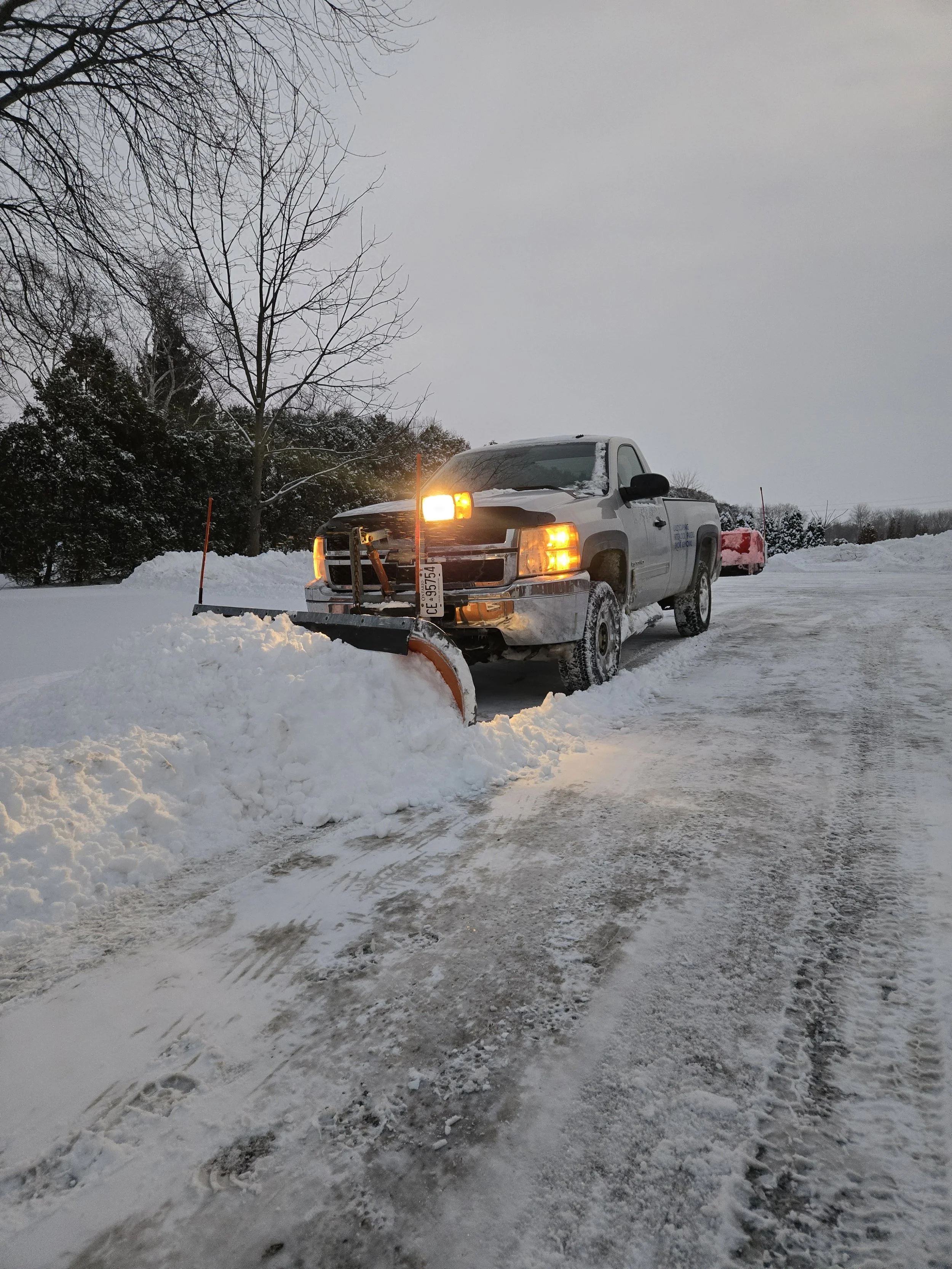 Red snowplow tractor clearing snow on a snowy day with houses and trees in the background.