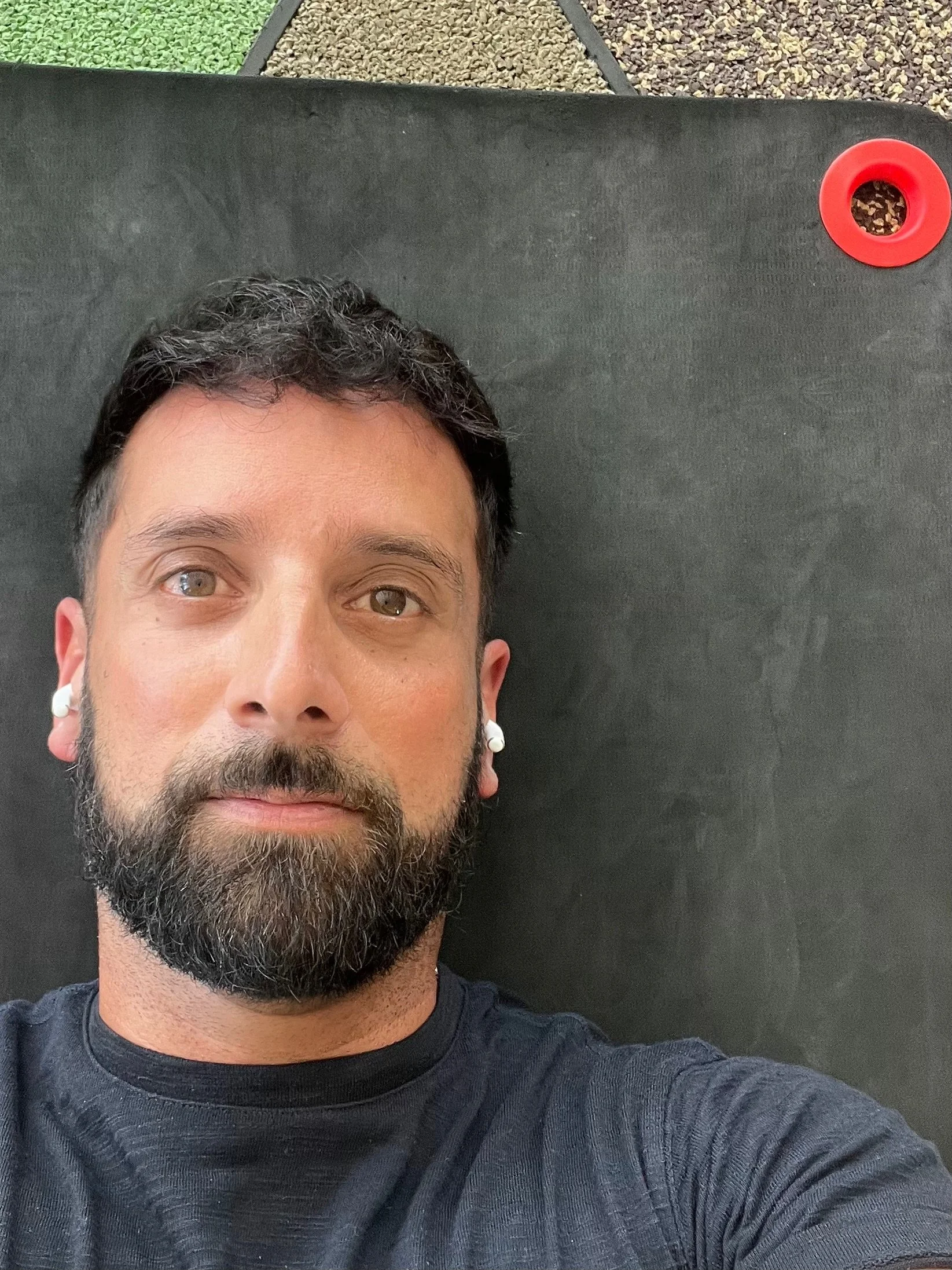 A man with a beard and short dark hair lying on a black exercise mat, wearing white earbuds, looking at the camera, with green artificial grass and gravel visible in the background.