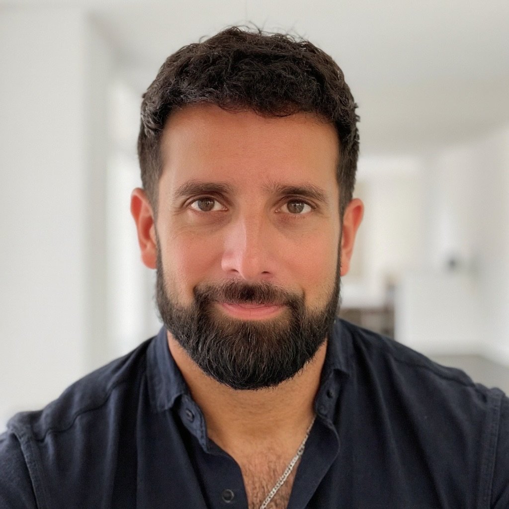 A portrait of a man with dark hair, a beard, wearing a dark shirt, looking at the camera with a slight smile in a well-lit room.