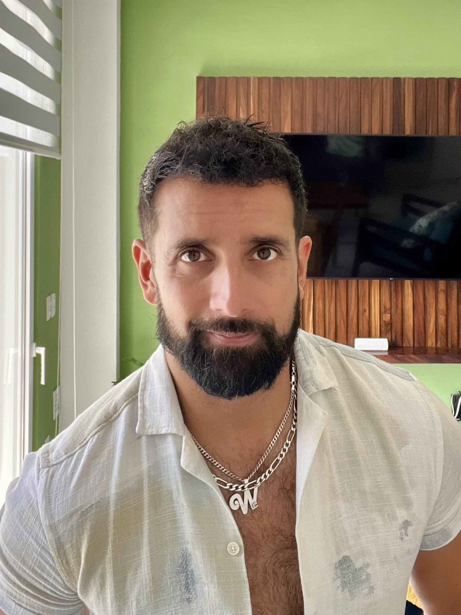 A man with dark hair and a beard wearing a beige shirt and silver chain necklace with a 'LOVE' pendant, inside a modern room with green walls and a wooden entertainment center with a large TV.