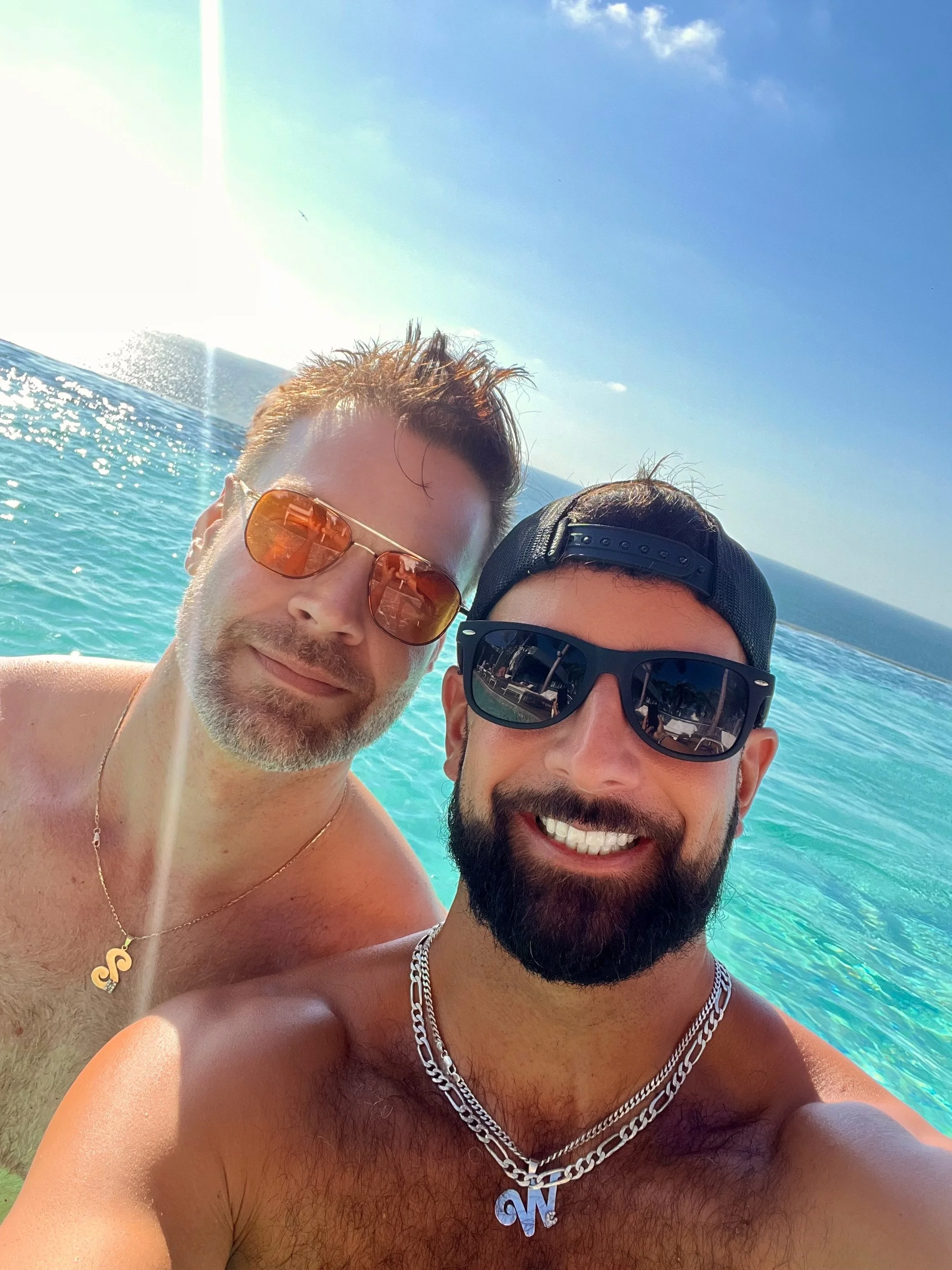 Two men are taking a selfie in the ocean on a sunny day, both wearing sunglasses and smiling.
