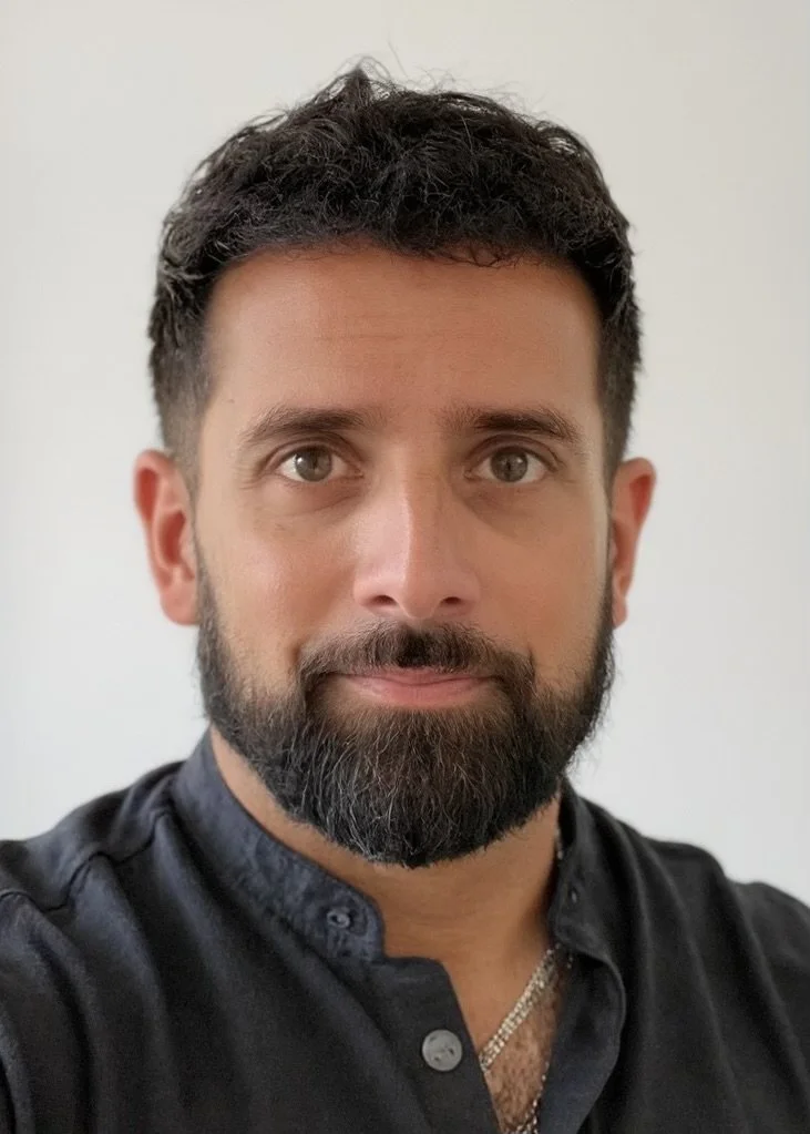 A close-up portrait of a man with dark, curly hair, a full beard, and brown eyes, wearing a black button-up shirt with a chain necklace visible, against a plain white background.