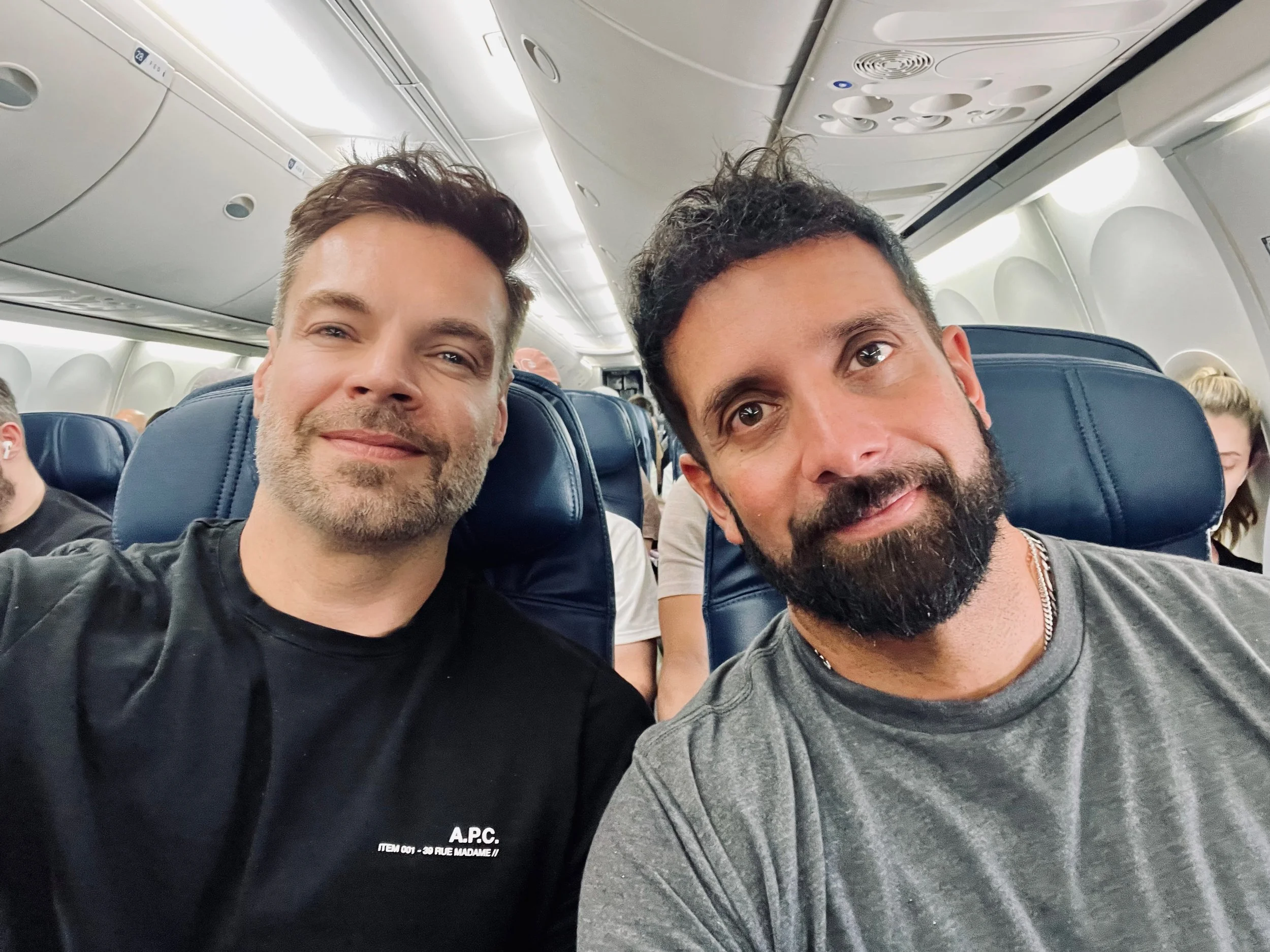 Two men sitting next to each other on an airplane, taking a selfie. They are smiling, with one wearing a black shirt and the other a gray shirt. The airplane cabin is visible in the background, with other passengers seated.