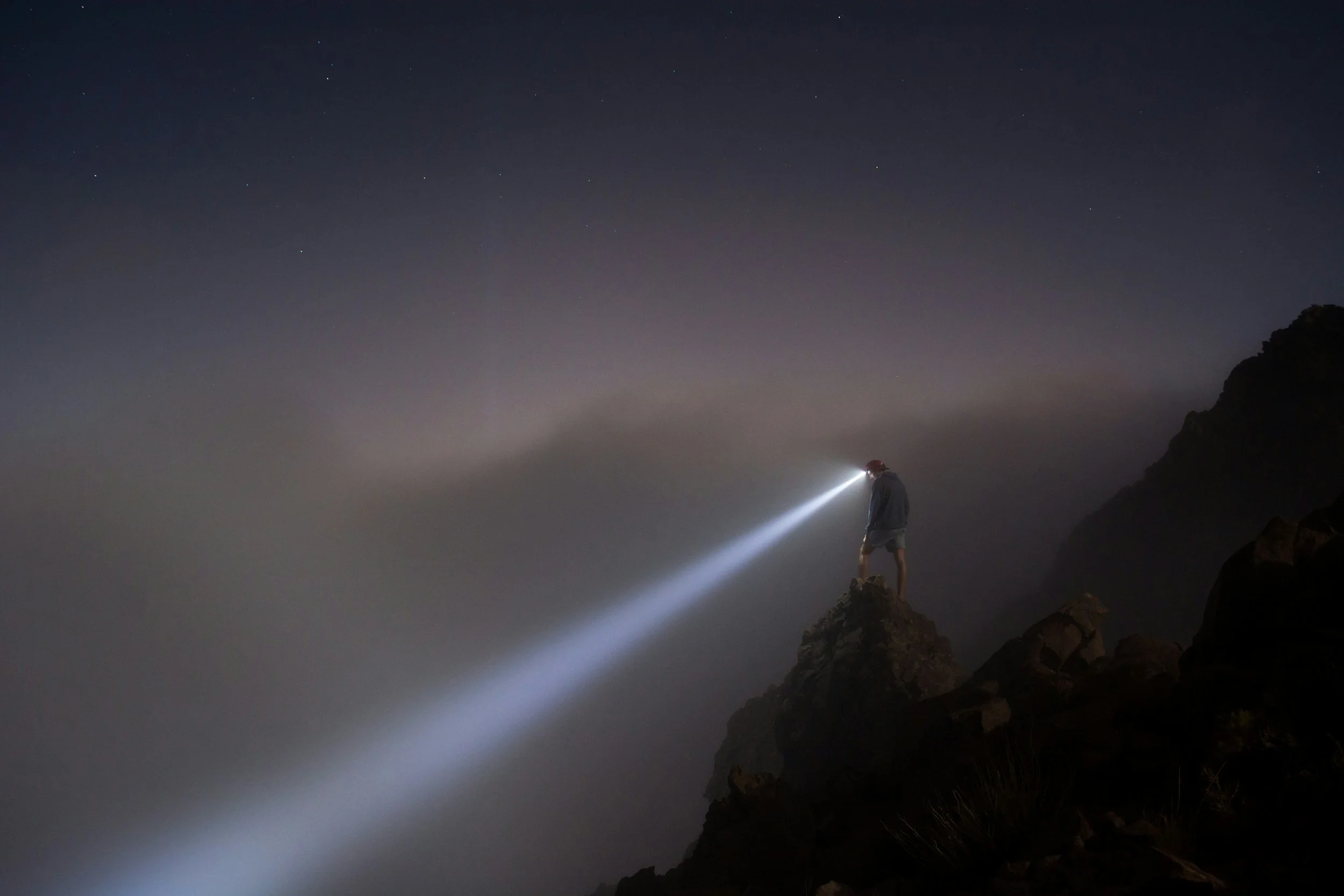 Person standing on a rocky cliff at night using a headlamp to illuminate the way, with a starry sky in the background.