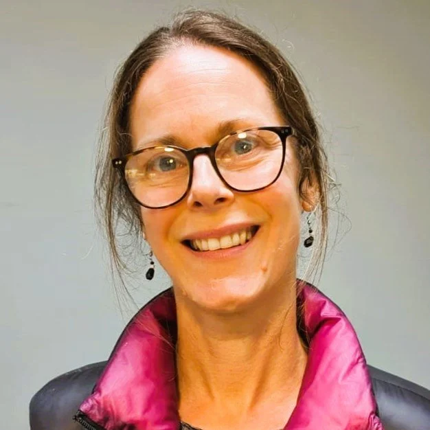 Close-up of a smiling woman with black hair, wearing a blue top, in a green indoor garden or conservatory.