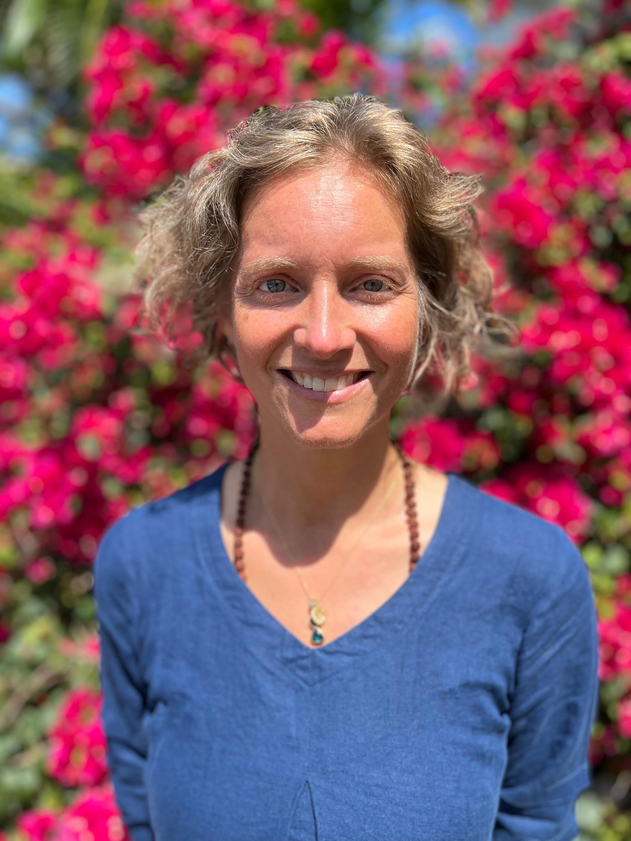 A man with curly dark hair, glasses, and a beard, wearing a dark green sweater and a white shirt, looking to the side with a slight smile, in a bright room with a plant in the background.