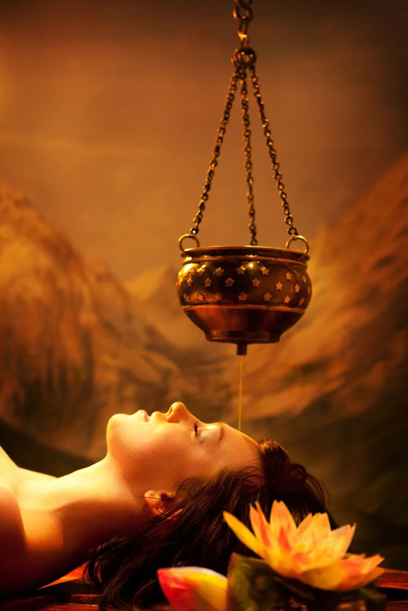 A glass teacup filled with herbal tea containing yellow, purple, and dark herbs, placed on a wooden surface. Around the cup are loose dried herbs and flower petals, with green fern leaves and white flowers nearby.