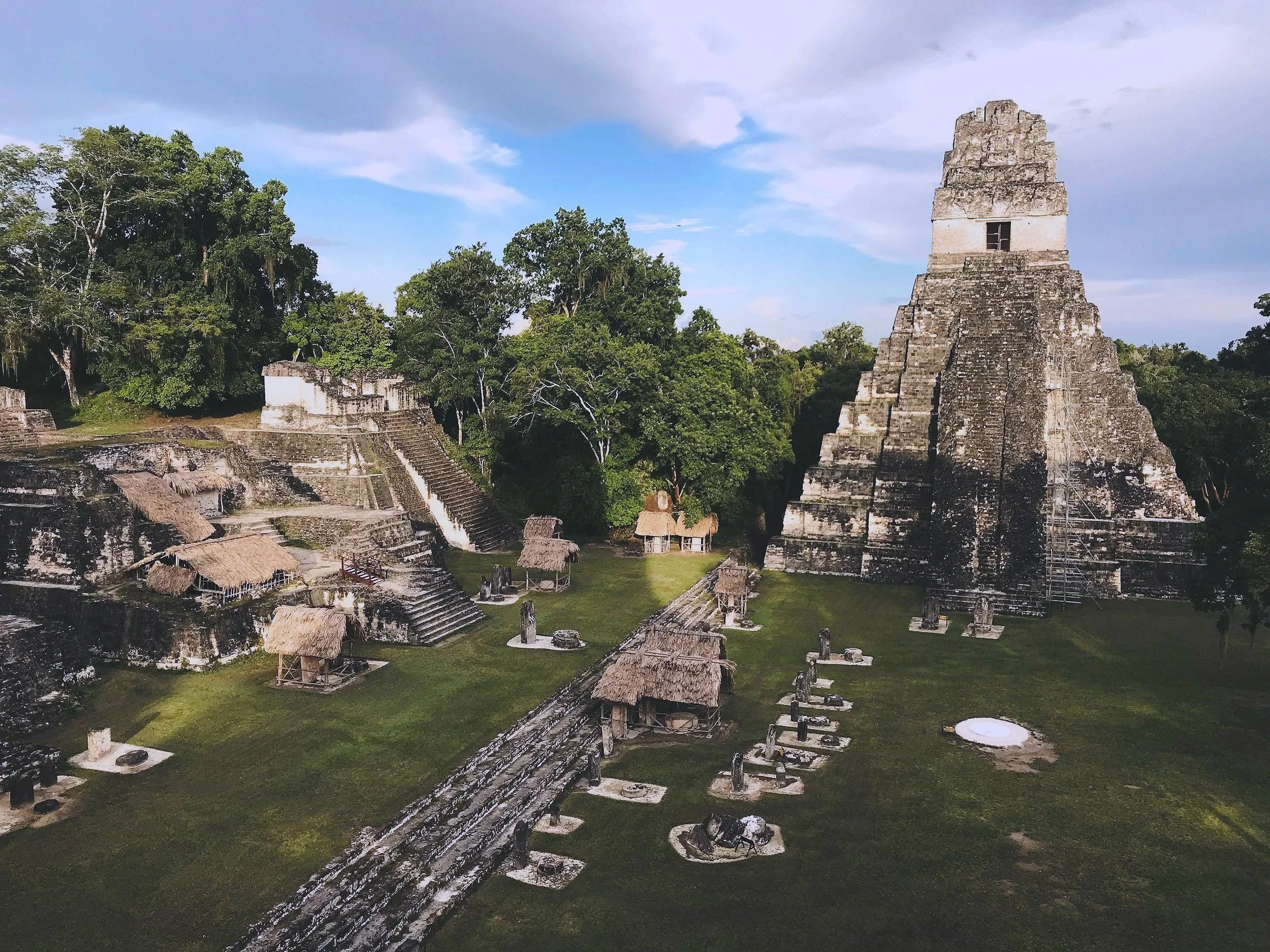 Ancient Mayan pyramid and temple ruins surrounded by green trees, with small thatched roof structures and statues in the grassy area in front.