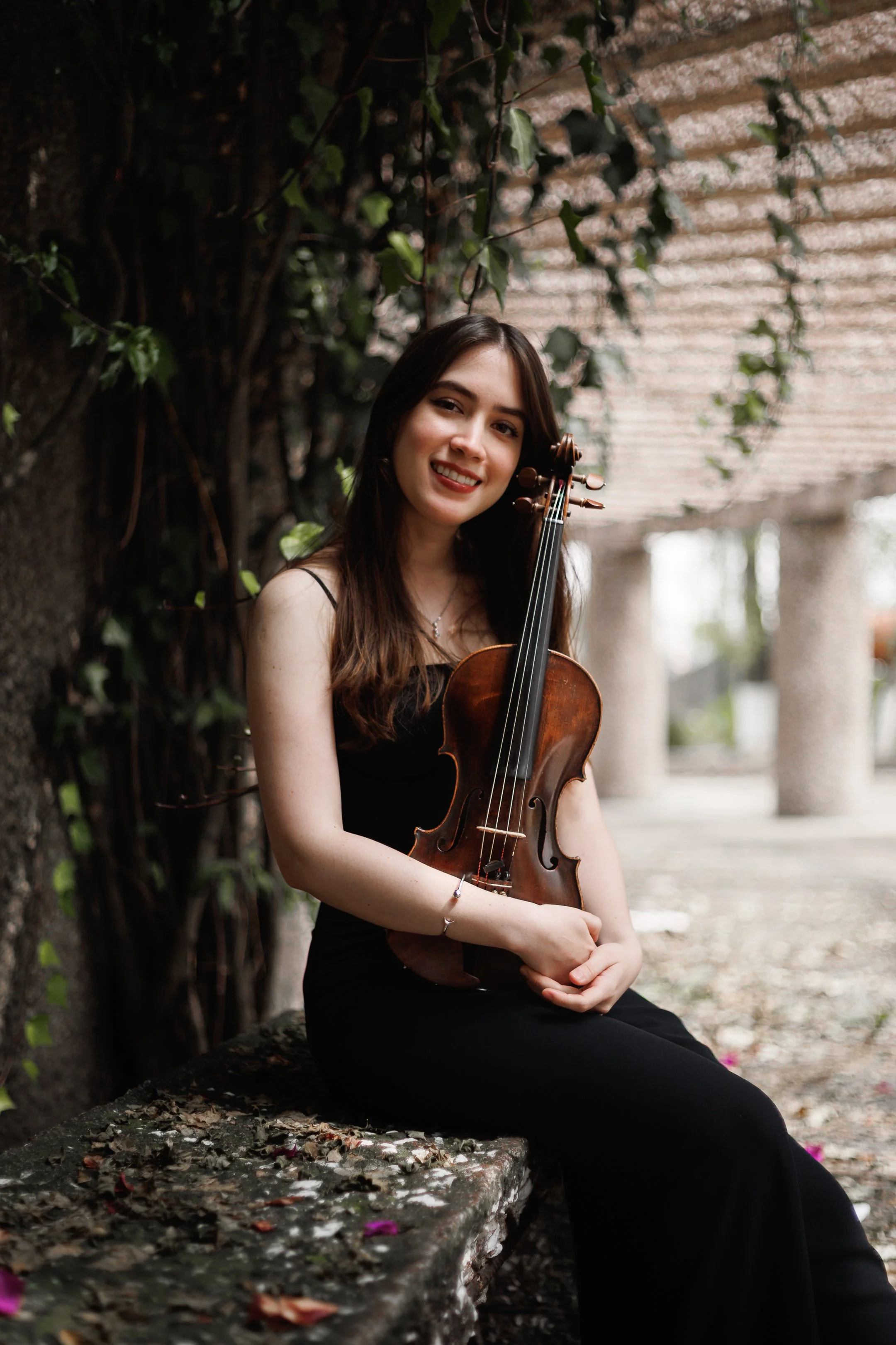 Joven mujer con cabello largo y oscuro, sonriendo, sosteniendo un violín, sentada en un banco de piedra en un lugar al aire libre con paredes cubiertas de hiedra y columnas de piedra al fondo.