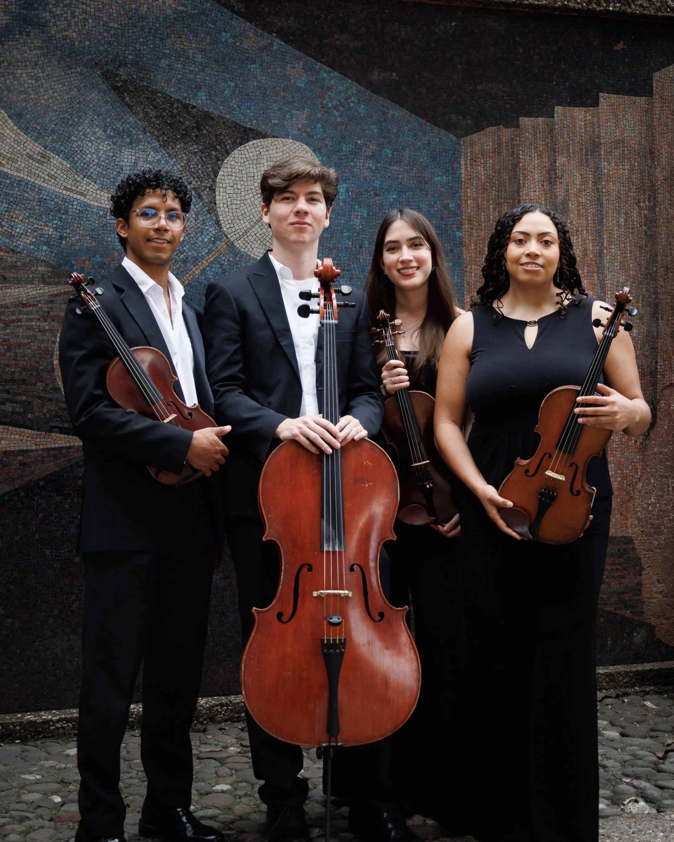 Cuatro jóvenes músicos con instrumentos de cuerda, dos viranolas y dos violines, posando frente a un mural de mosaico con un diseño abstracto en tonos azules y marrones.