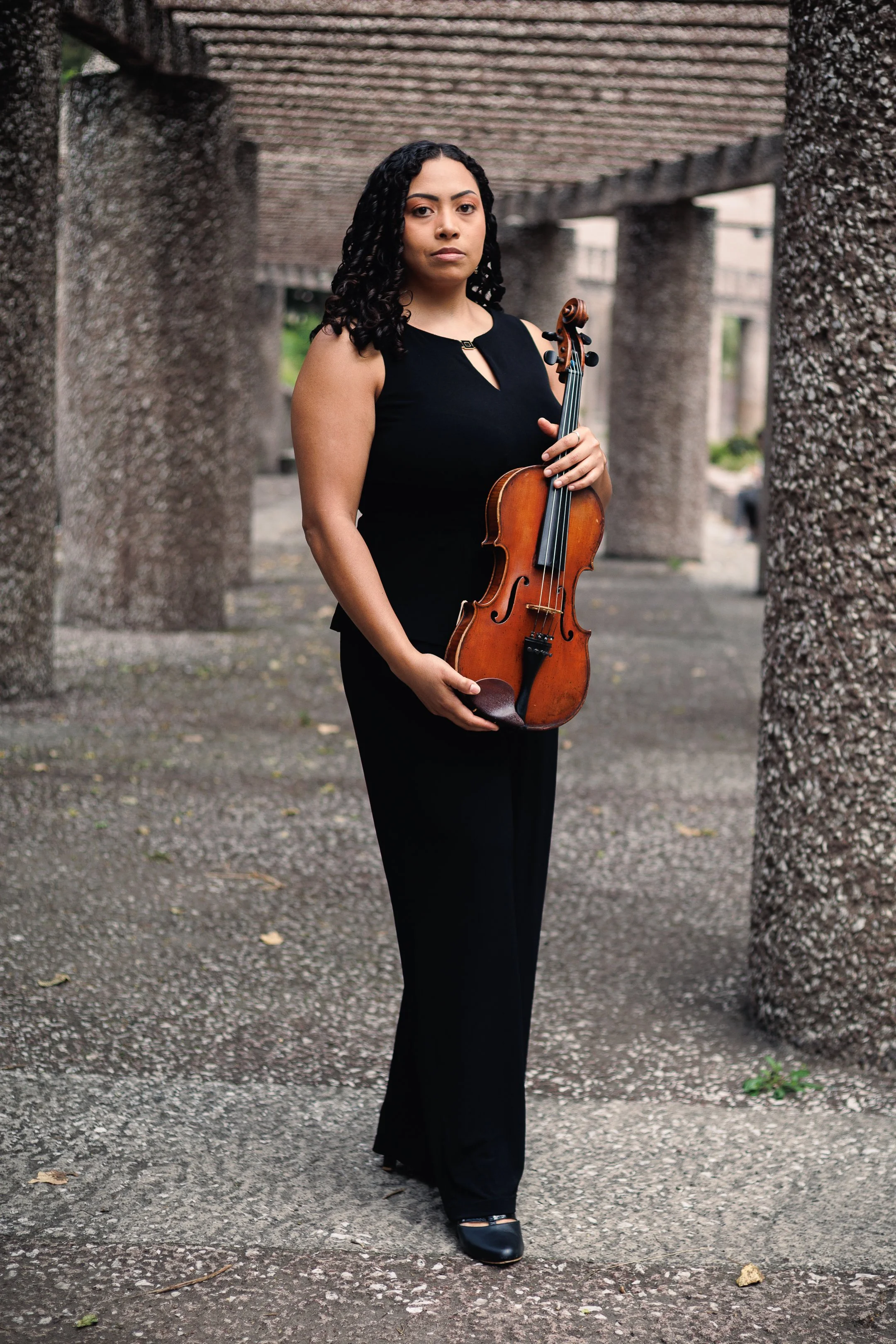 Mujer con cabello rizado, vestida con un vestido negro, sostiene un violín, caminando por un pasillo con columnas de concreto en un entorno exterior.