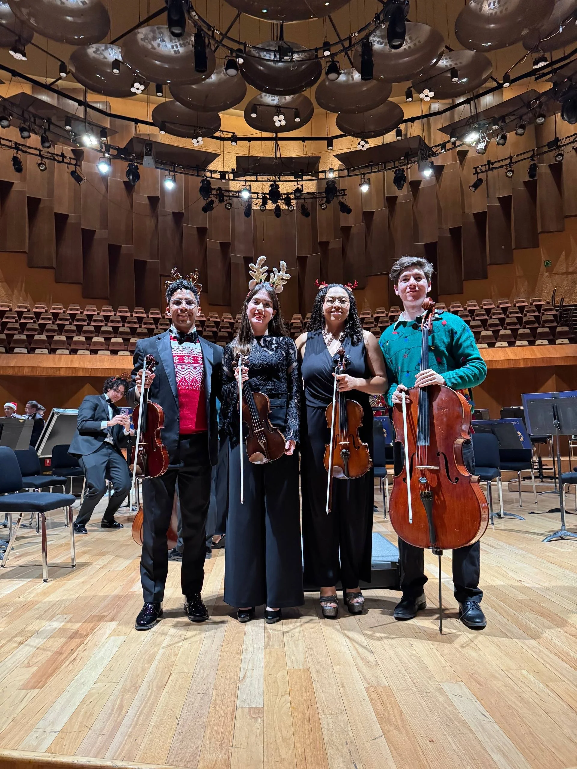 Cuatro músicos de orquesta sosteniendo violines y un chelo en un escenario de concierto, con decoraciones navideñas y miembros del público y personal en el fondo.