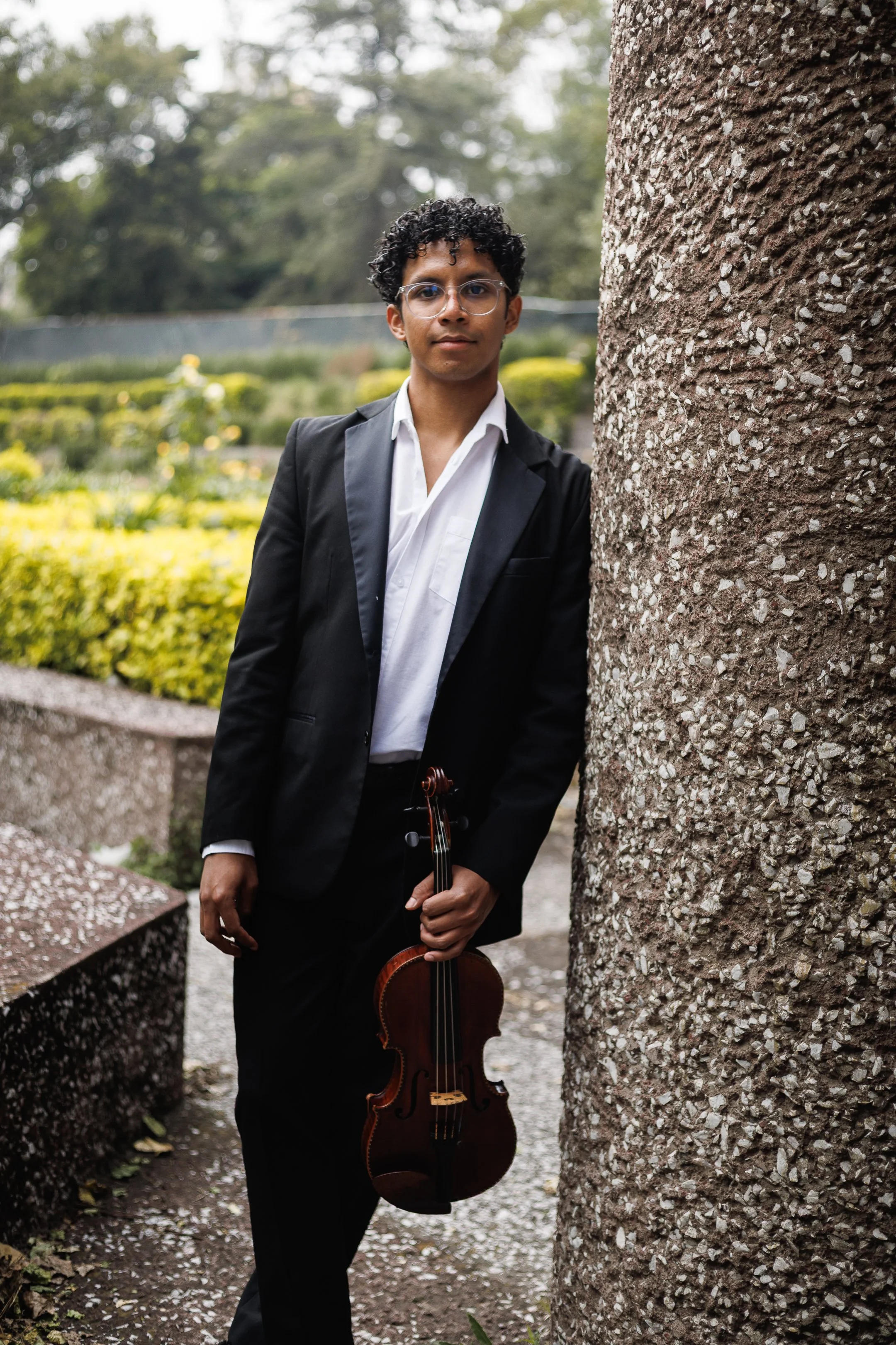Joven hombre con gafas, vestido con traje y sosteniendo un violín, posando de pie junto a un muro de piedra en un parque con áreas verdes y árboles al fondo.