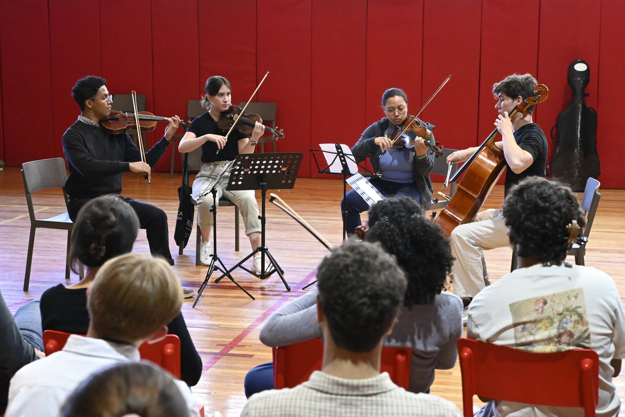 Cuatro músicos tocando violín y viola en un escenario rojo, con audiencia observando.