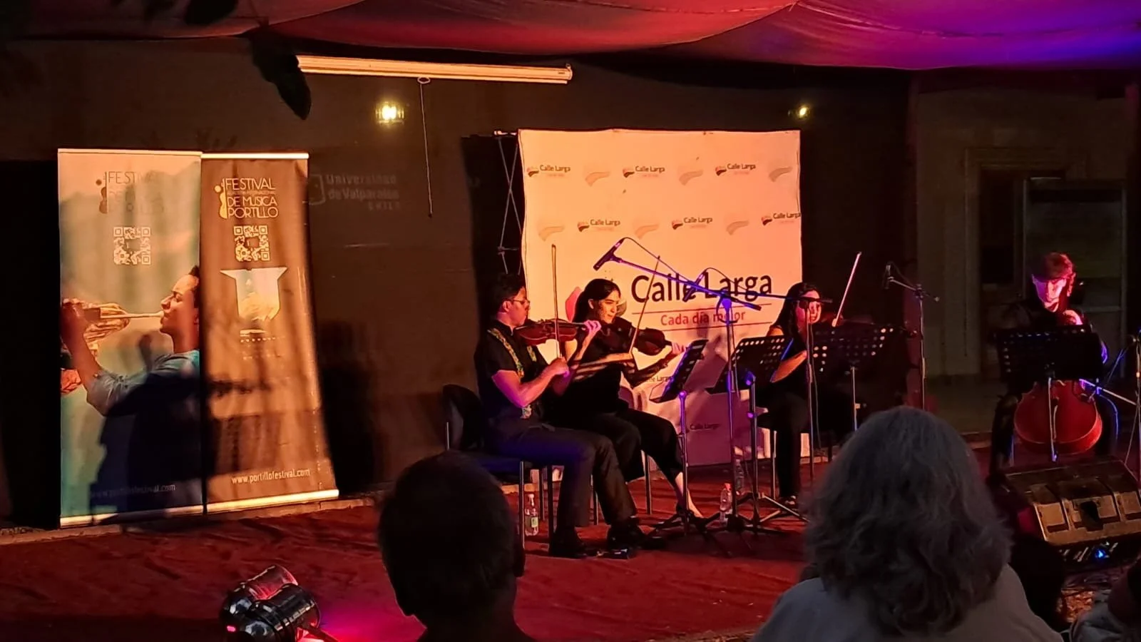 Cuatro músicos tocando en un escenario, acompañados por micrófonos y partituras, con banners de fondo que promocionan un festival de música y una entidad llamada Calle Larga.