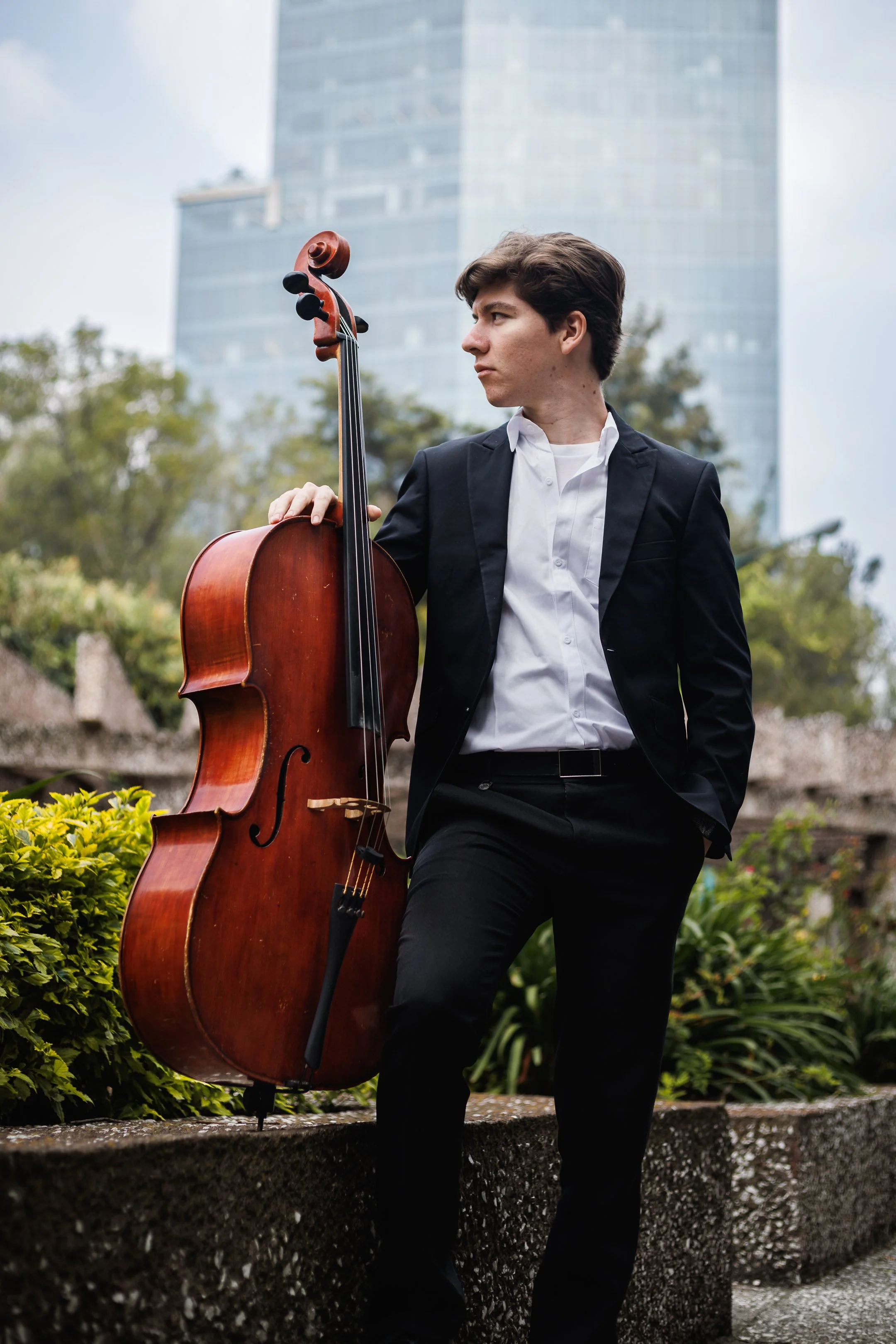 Joven hombre con cabello rizado y vestir formal con saco negro y camisa blanca, sosteniendo un violonchelo en un espacio exterior, con edificios modernos y árboles en el fondo.