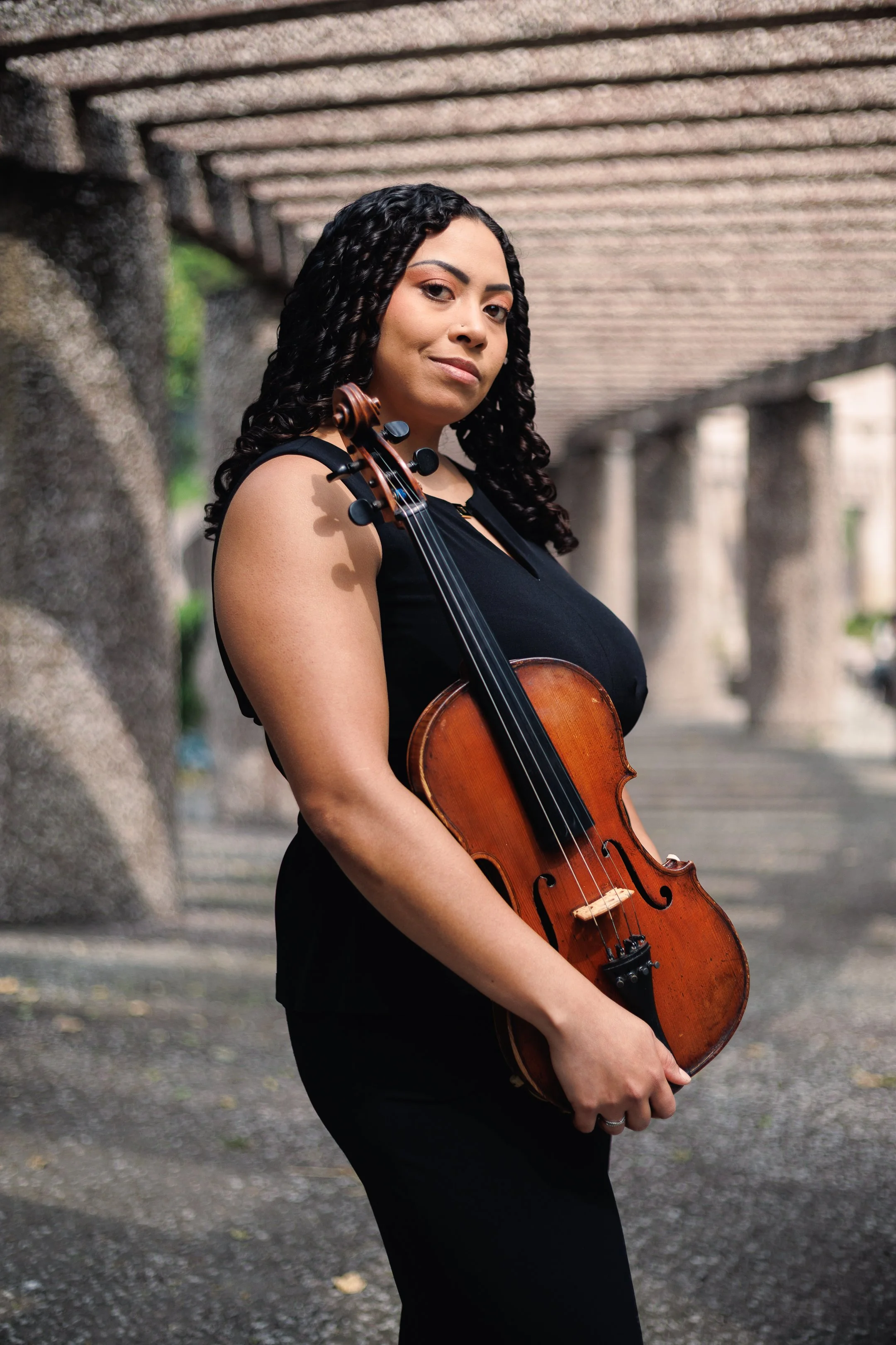 Mujer con cabello rizado oscuro, vestida con ropa negra, sostiene un violín en un entorno urbano con estructura de cemento en el fondo.