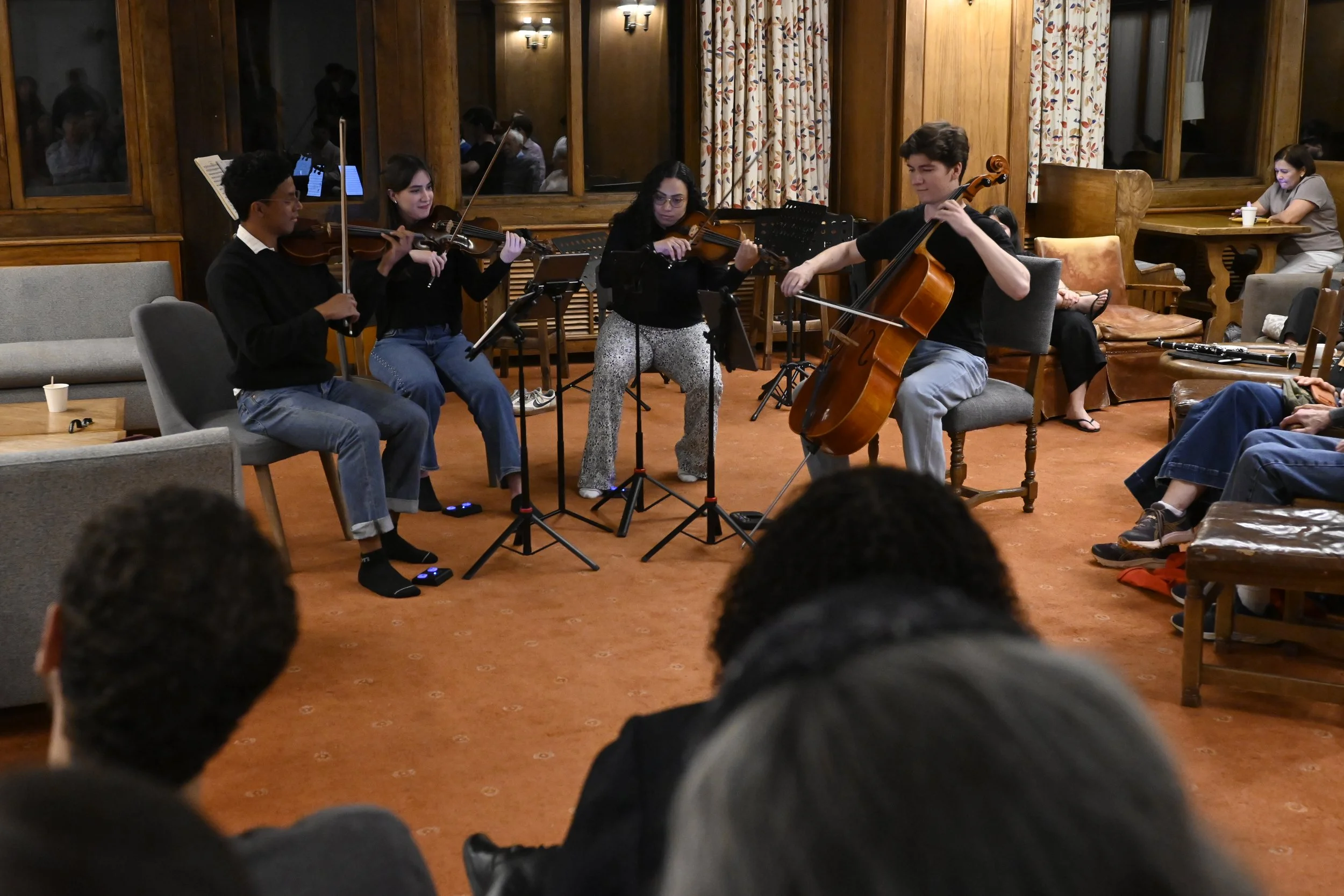 Un grupo de músicos tocando violín y violonchelo en una sala con decoración de madera y cortinas con follaje.