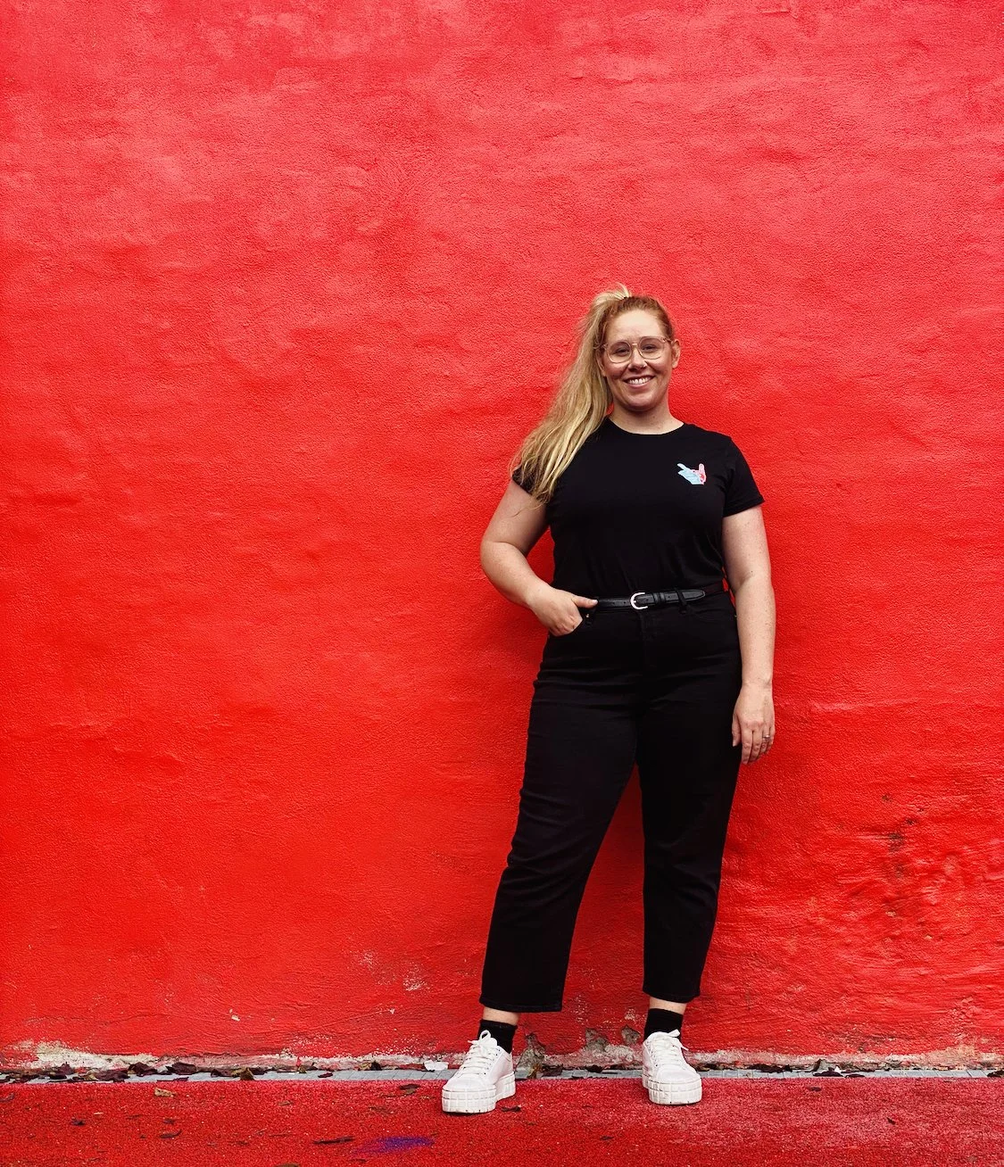 A woman standing against a bright red wall, smiling, wearing glasses, black t-shirt with a small colorful graphic, black pants with a belt, and white sneakers.