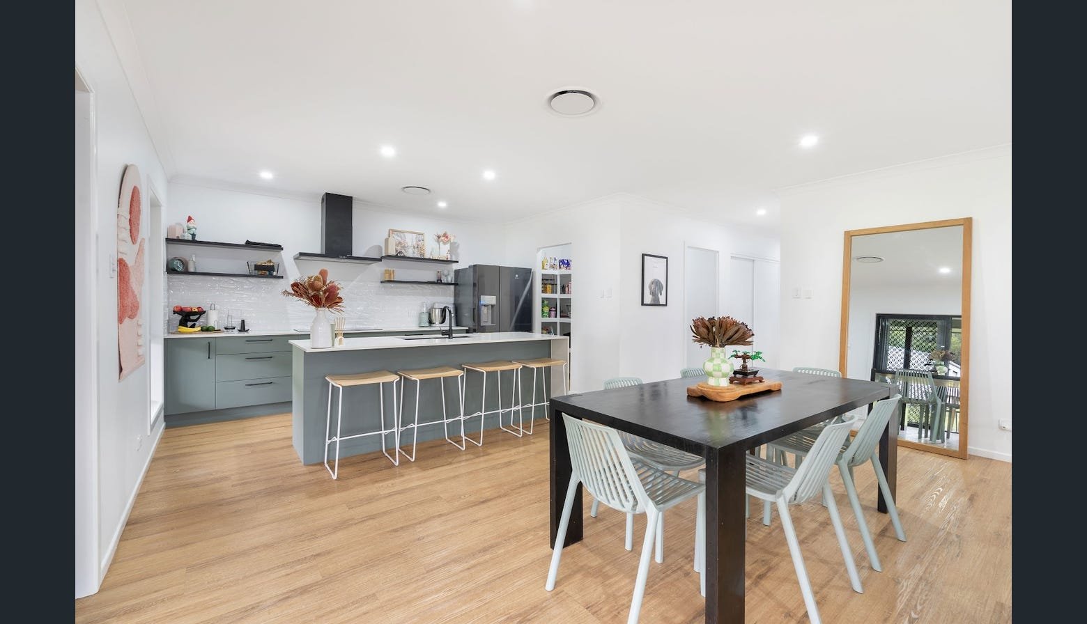 Modern open-concept kitchen and dining area with wooden flooring, gray kitchen cabinets, a large island with bar stools, a black dining table with white chairs, and large mirrors reflecting the room.