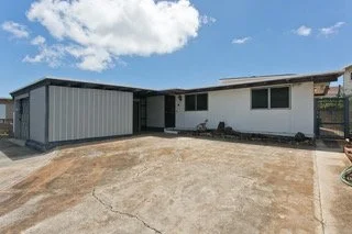 A single-story house with a gray metal shed on the left, a white exterior, and a large concrete driveway in front under a partly cloudy sky.
