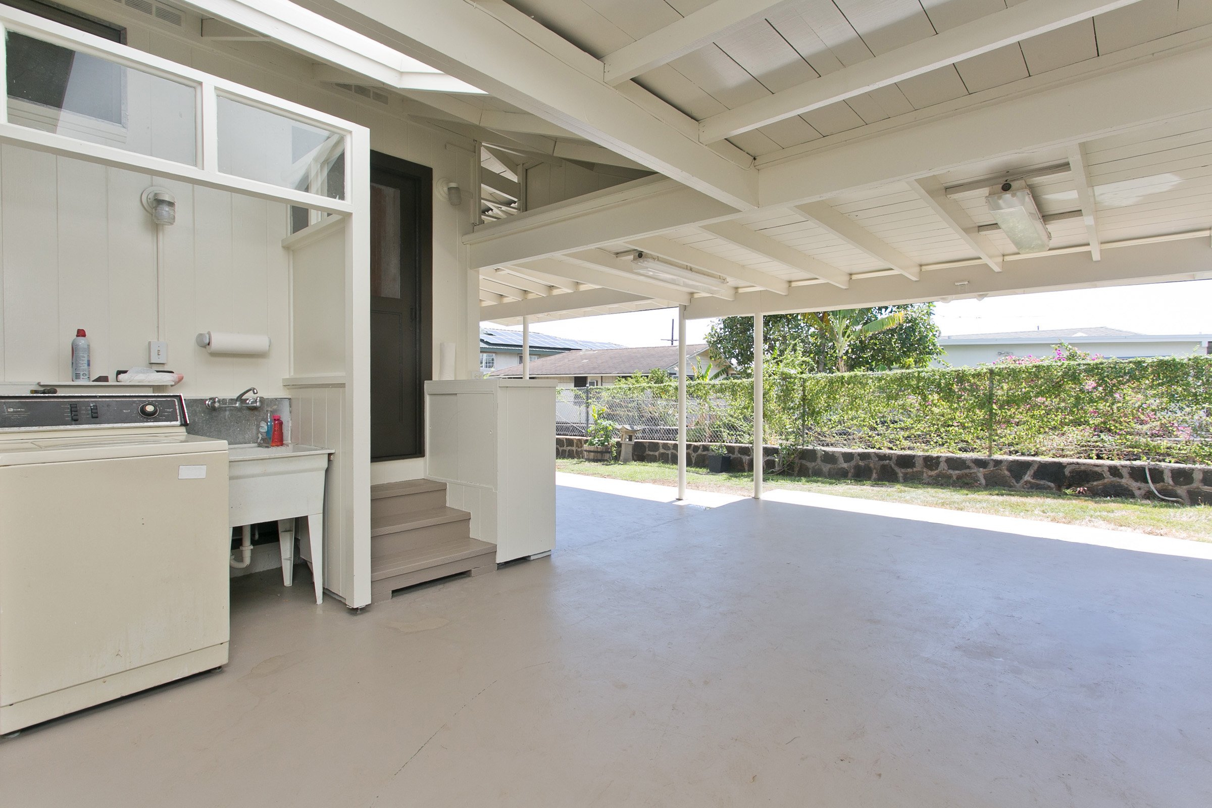 Empty covered patio with a laundry sink, washing machine, and steps leading to an interior door, overlooking a backyard with a stone border and green bushes.