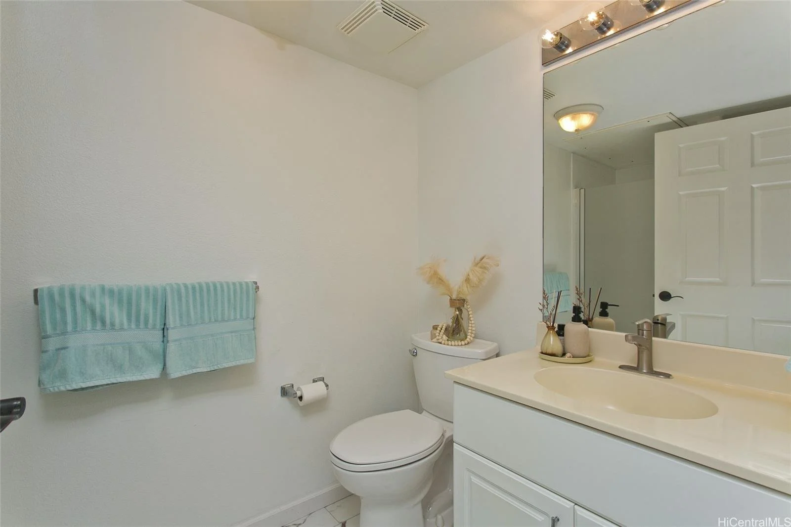 A small bathroom with a white toilet, a white vanity with a sink, and a large mirror. The decor includes a decorative vase with dried grasses and small figurines on the toilet tank, and a soap dispenser and tray on the sink. There is a light fixture