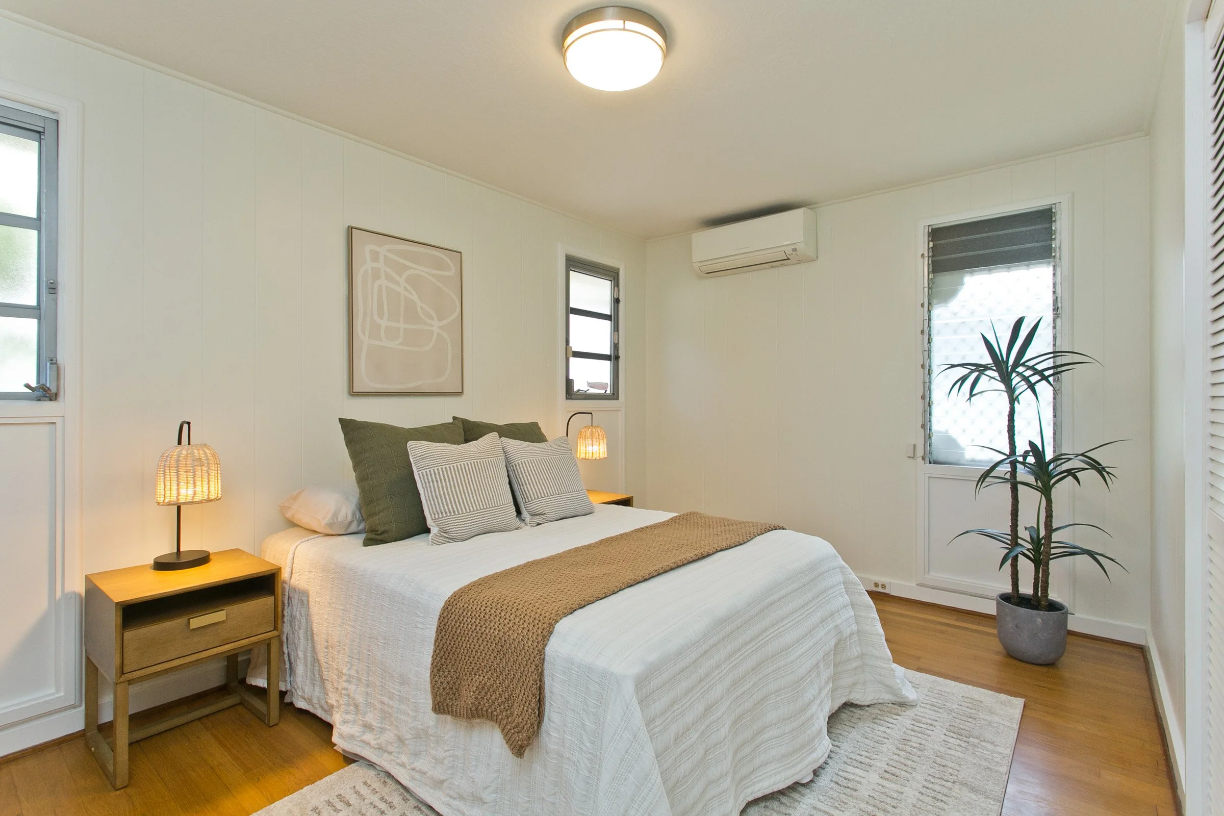 Bright bedroom with white walls, wooden floor, and a bed with white bedding, green and patterned pillows, and a brown blanket. Two wooden nightstands with wicker lamps, a large potted plant in a gray pot, a wall-mounted air conditioner, and a framed