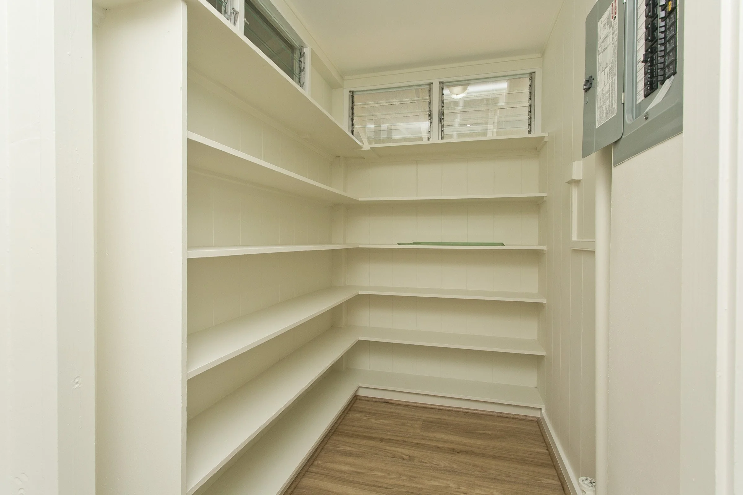 Empty beige pantry with multiple shelves, wood floor, and small windows at the top.