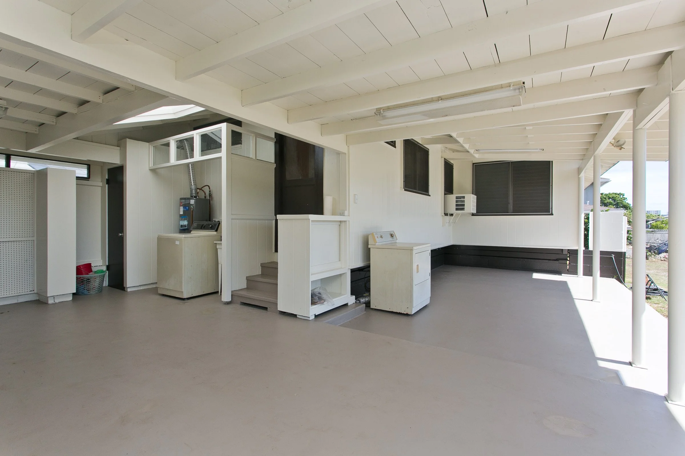 Empty covered outdoor patio area with white wooden beams, a white washing machine, an air conditioning unit, and windows with black shutters.