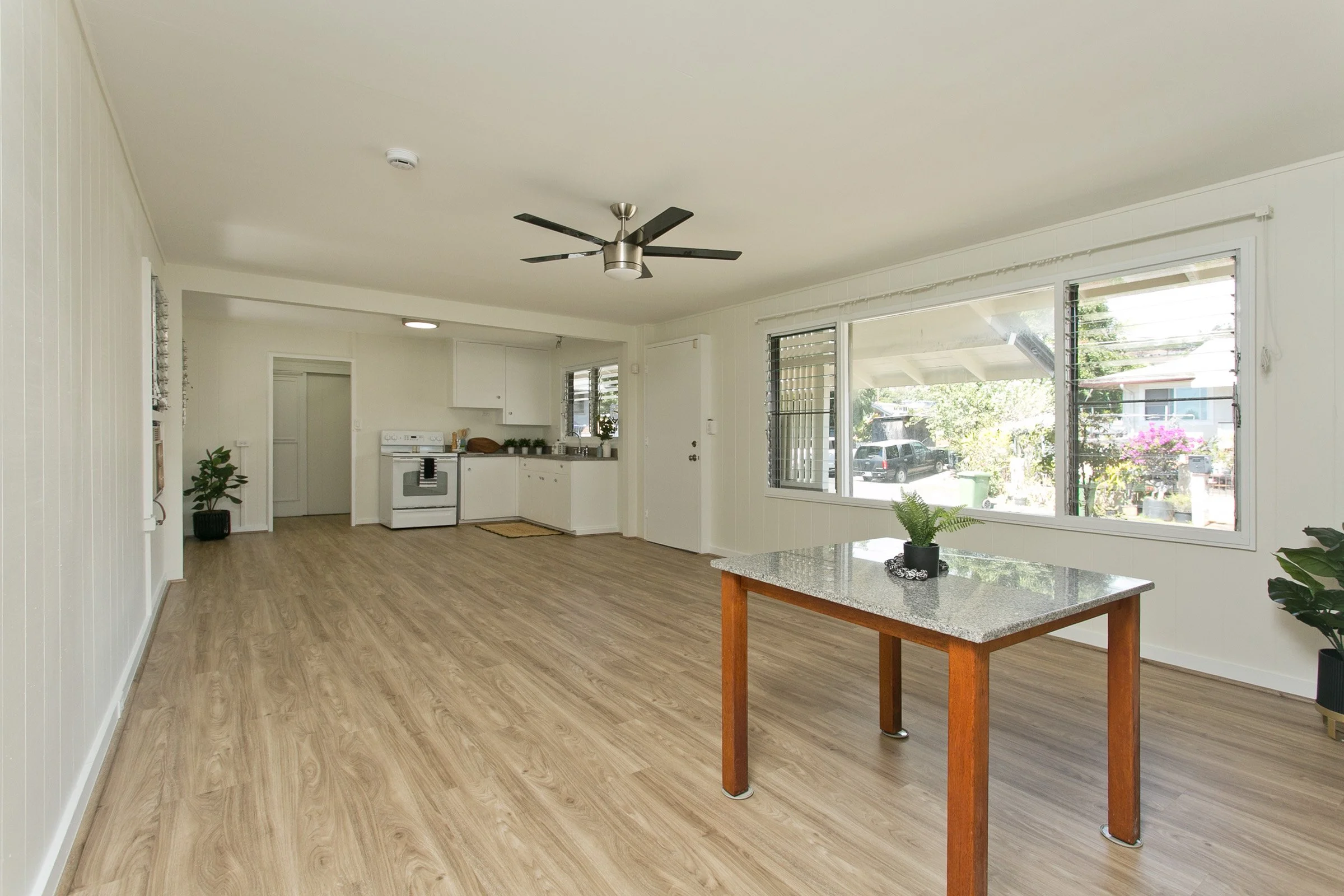 Open living space with wood-like flooring, white walls, large windows, a potted plant, a wooden table with a granite top, and a ceiling fan. A kitchen area with white cabinets and a stove is visible in the background.