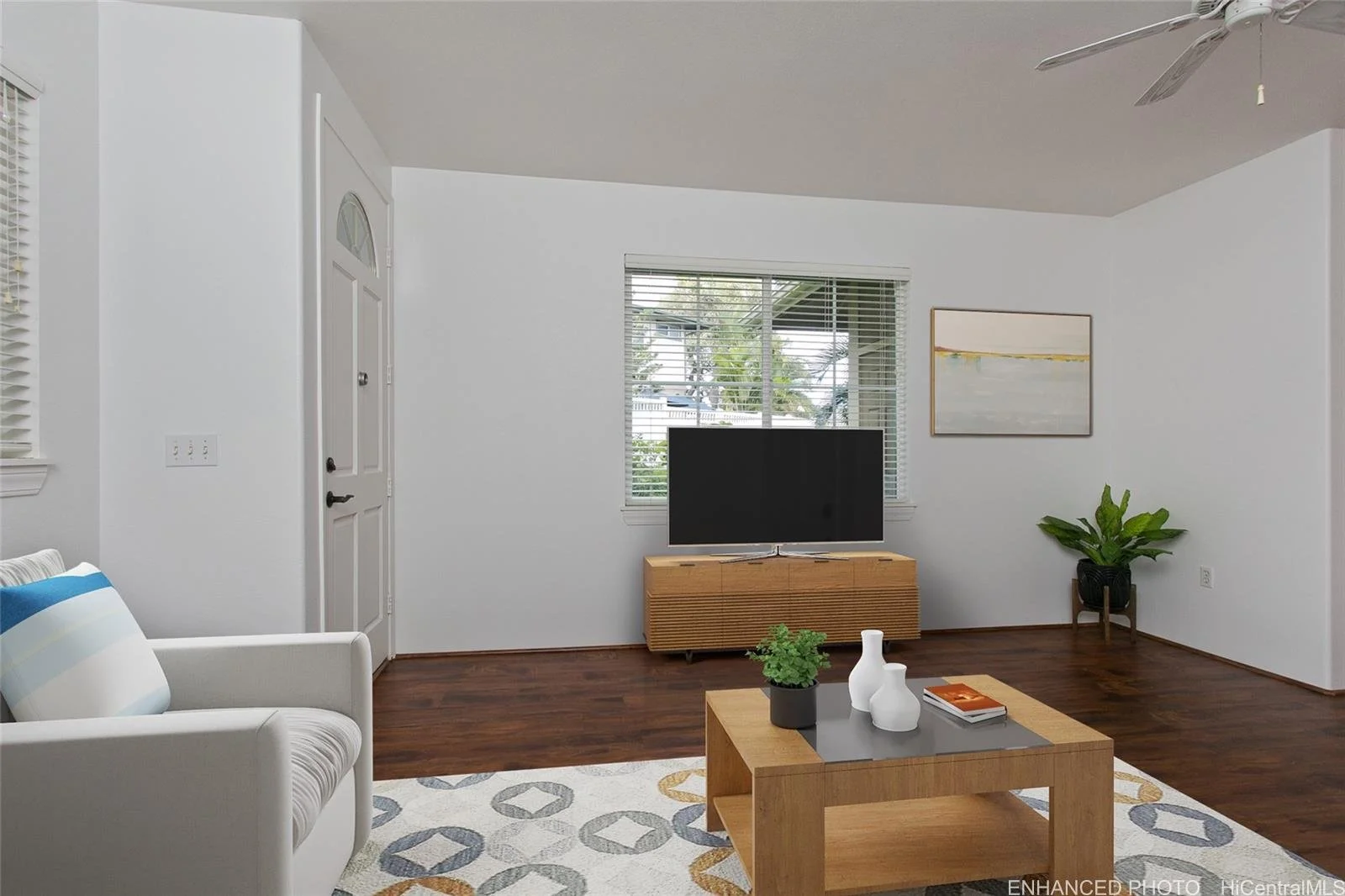 Living room with white walls, wooden flooring, a white armchair with a striped pillow, a wooden coffee table with decorative vases and a magazine, a TV on a wooden stand, a large window with blinds, a potted plant, a wall art piece, and a ceiling fan