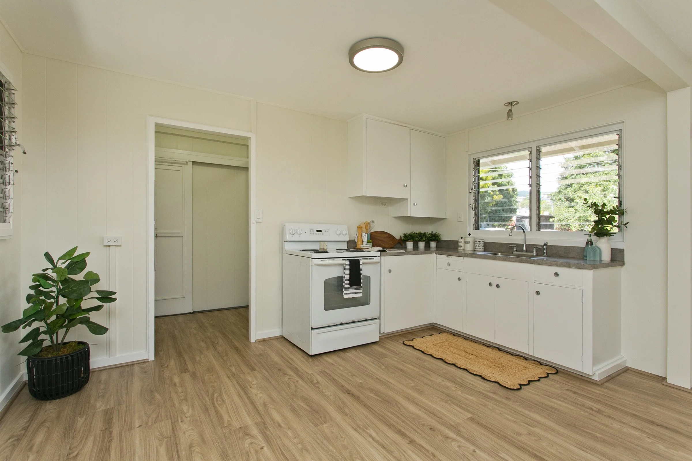 A simple, modern kitchen with white cabinets, a window with shutters, a white stove, a brown countertop, and a potted plant near a door. There is also a small rug on the light wood floor.