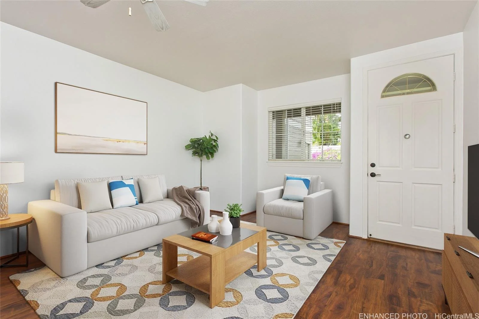 Bright living room with white sofa and armchair, wooden coffee table with decorative items, patterned area rug, wall art, window with blinds, potted plant, and white front door.