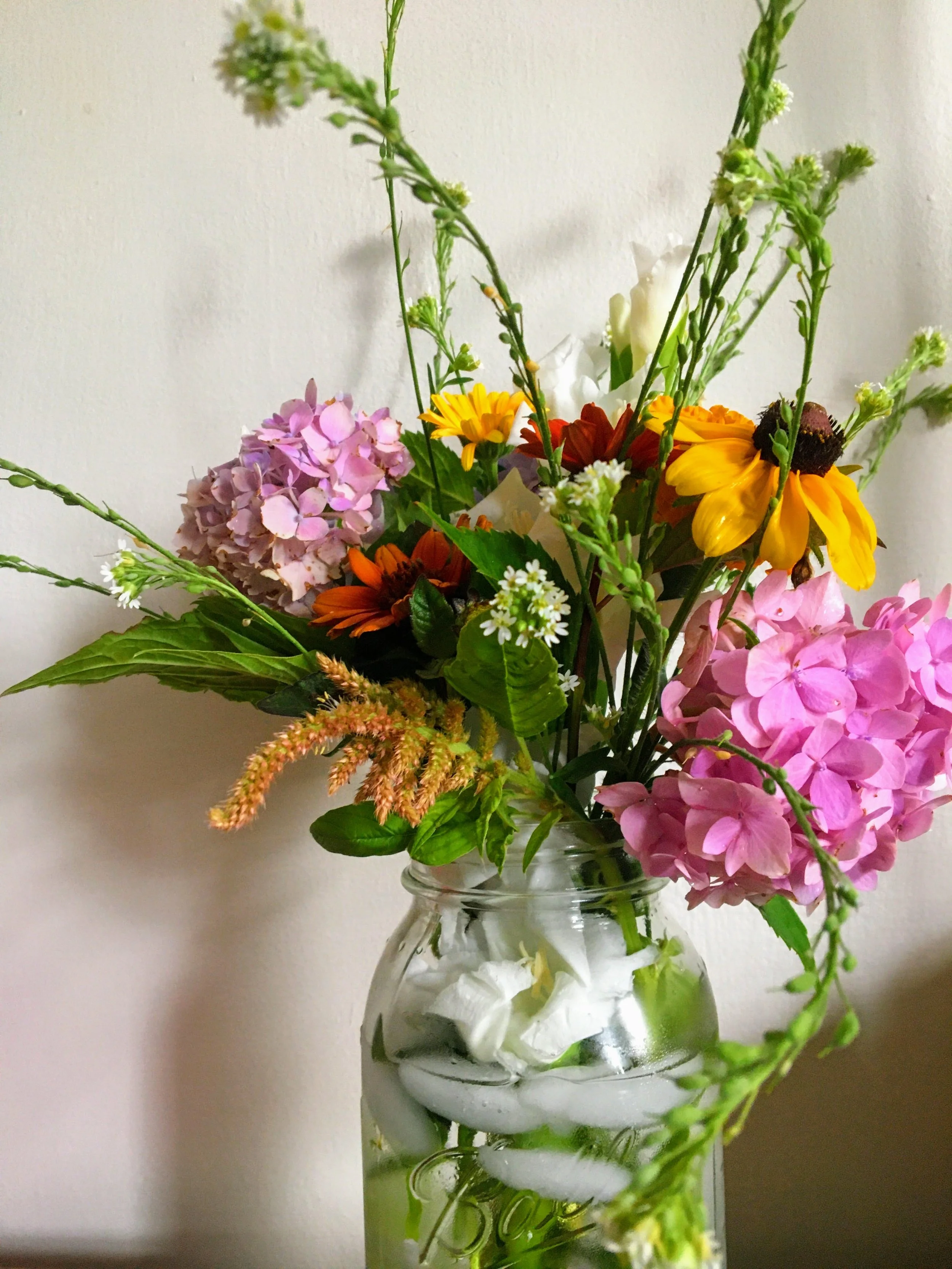 A glass jar filled with water and artificial flowers, including pink hydrangeas, yellow daisies, white blossoms, and green foliage.