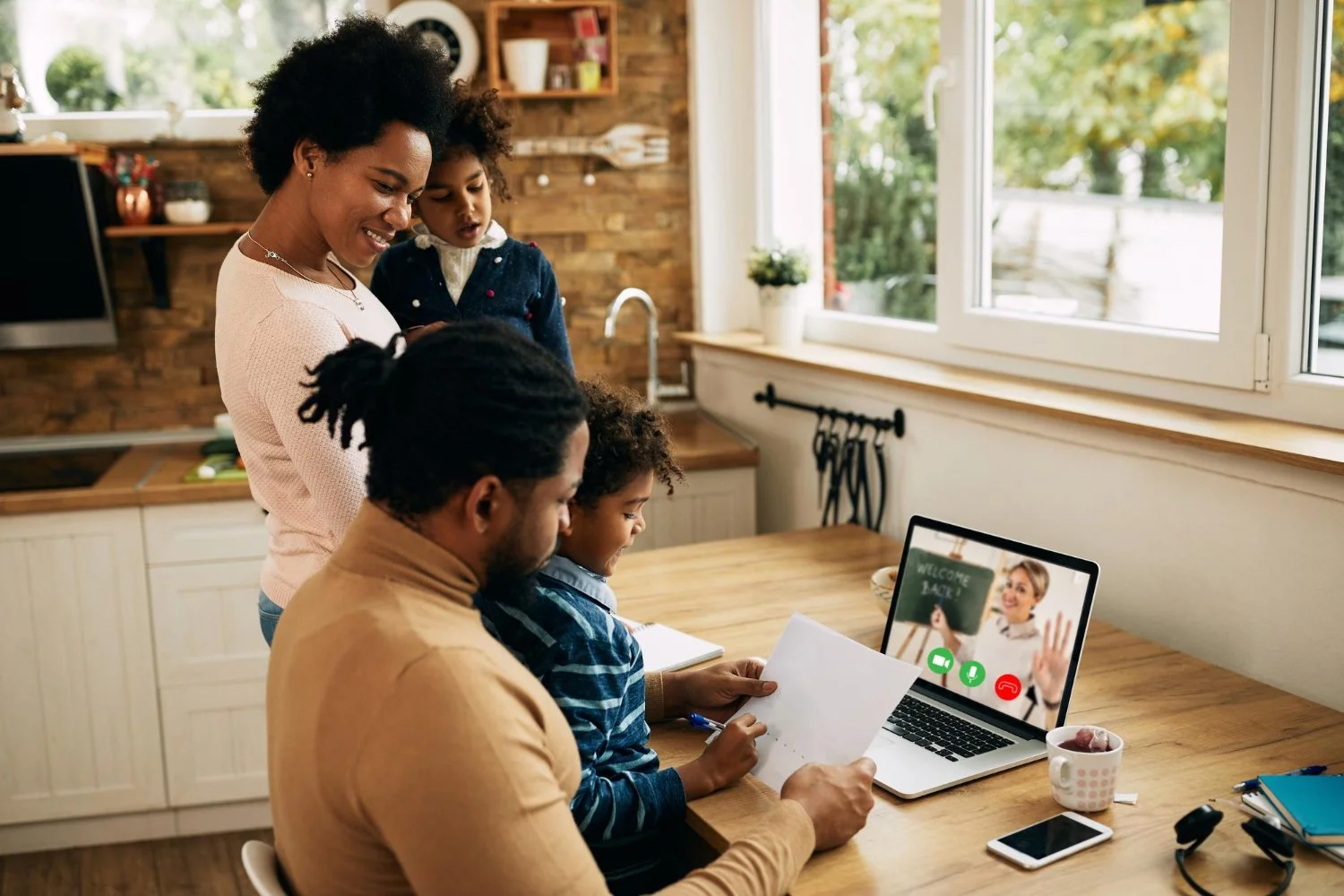 A family in a kitchen engaged in a video call on a laptop, with two children and a woman looking at papers, while a man is seated and using the laptop. The laptop screen shows a woman welcoming with a chalkboard that says "Welcome Back".