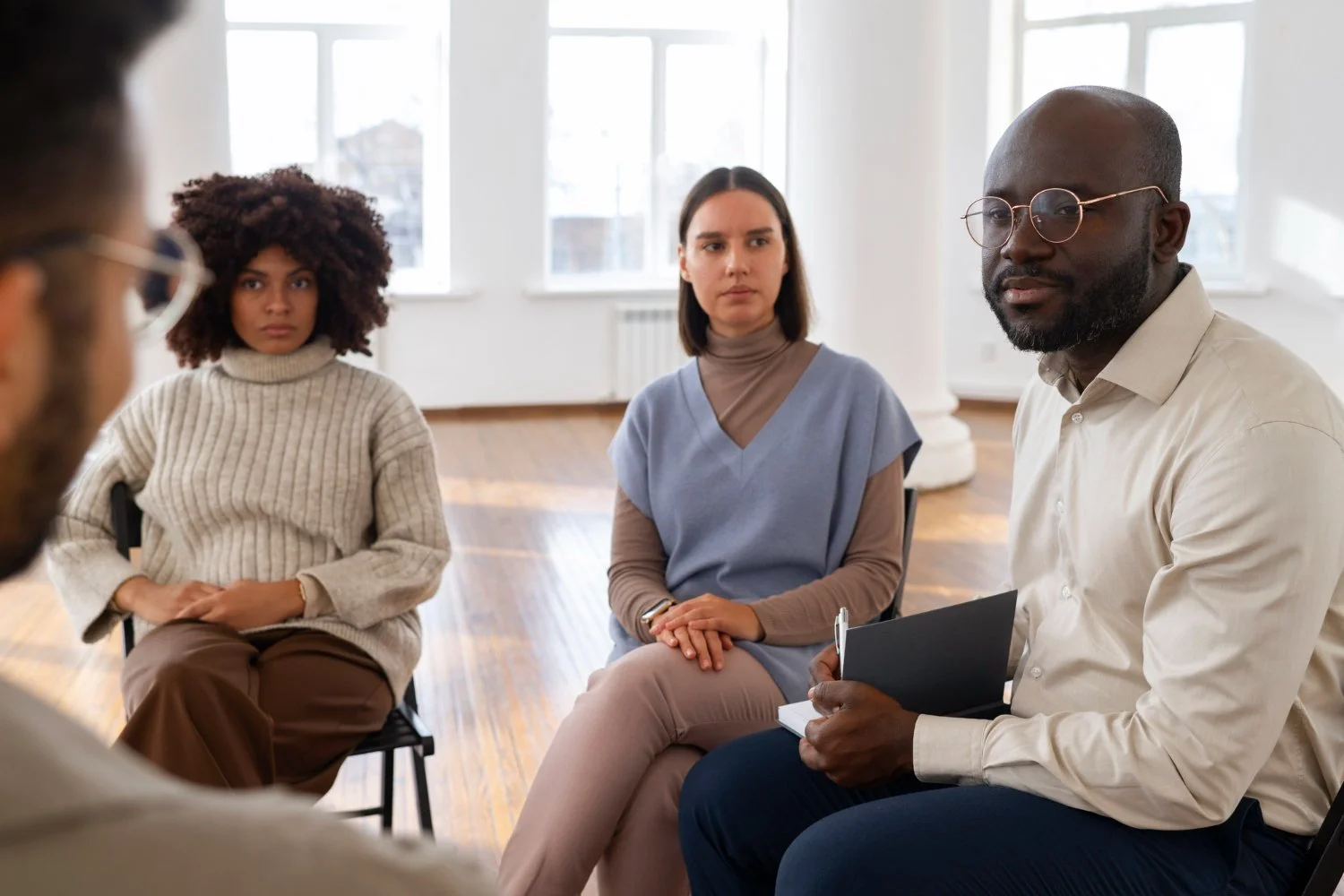 Four diverse adults sitting and listening during a discussion in a bright room with large windows.