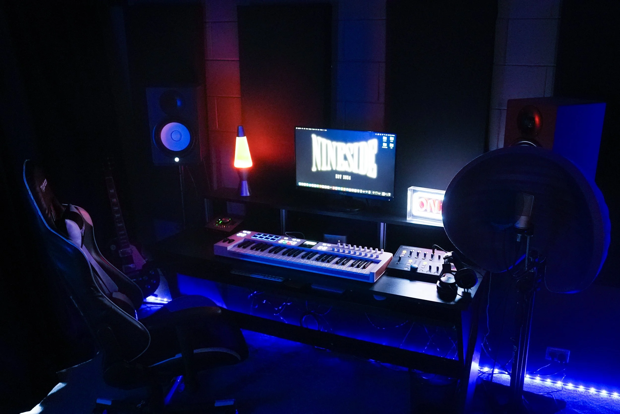A music studio with a computer monitor, keyboard, electronic music device, speakers, a lava lamp, and a guitar, illuminated by blue and red LED lights.