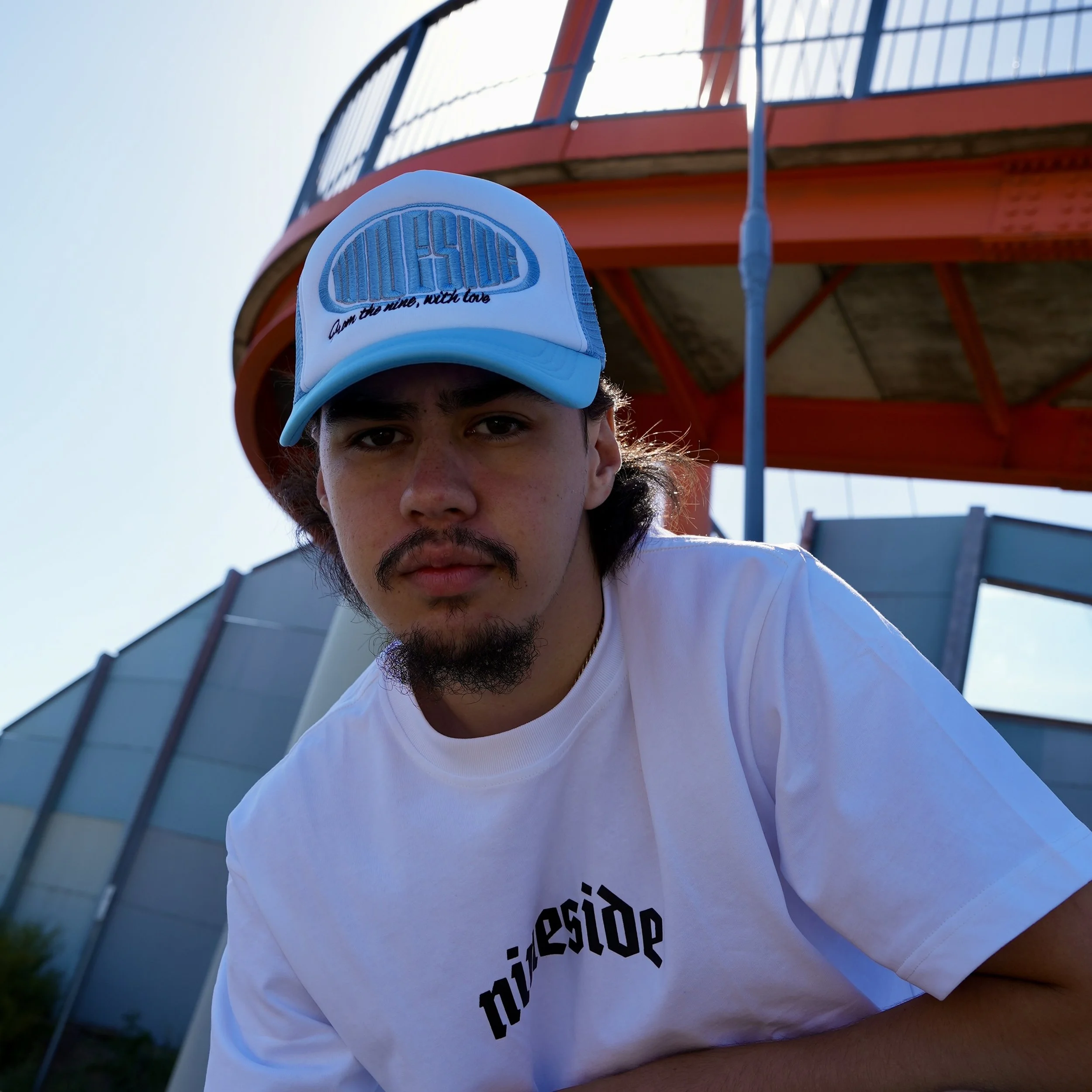 A young man with dark hair, a mustache, and a goatee, wearing a white t-shirt with black text and a blue cap, sitting outdoors near a red and blue structure.