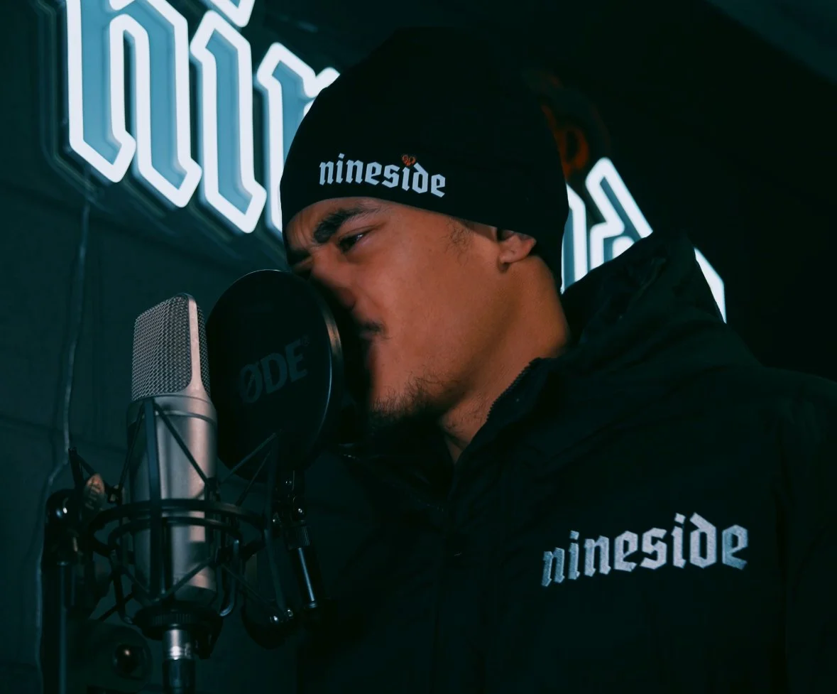A man wearing a black beanie with the word 'nineside' on it, singing into a microphone in a recording studio, with a neon sign behind him.