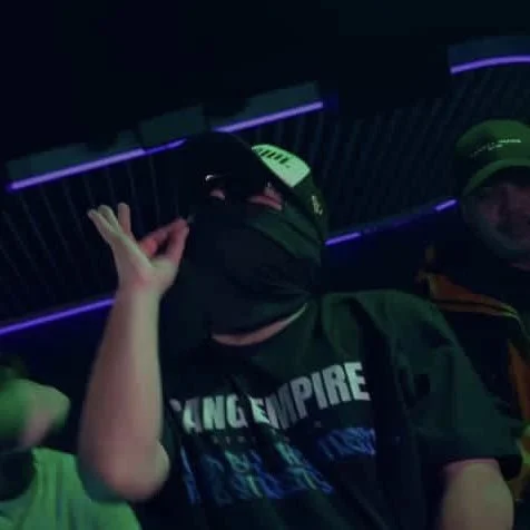 Three individuals in a dark indoor setting with neon purple lights, one person wearing a black hoodie, a cap, and headphones, making a peace sign with their hand.