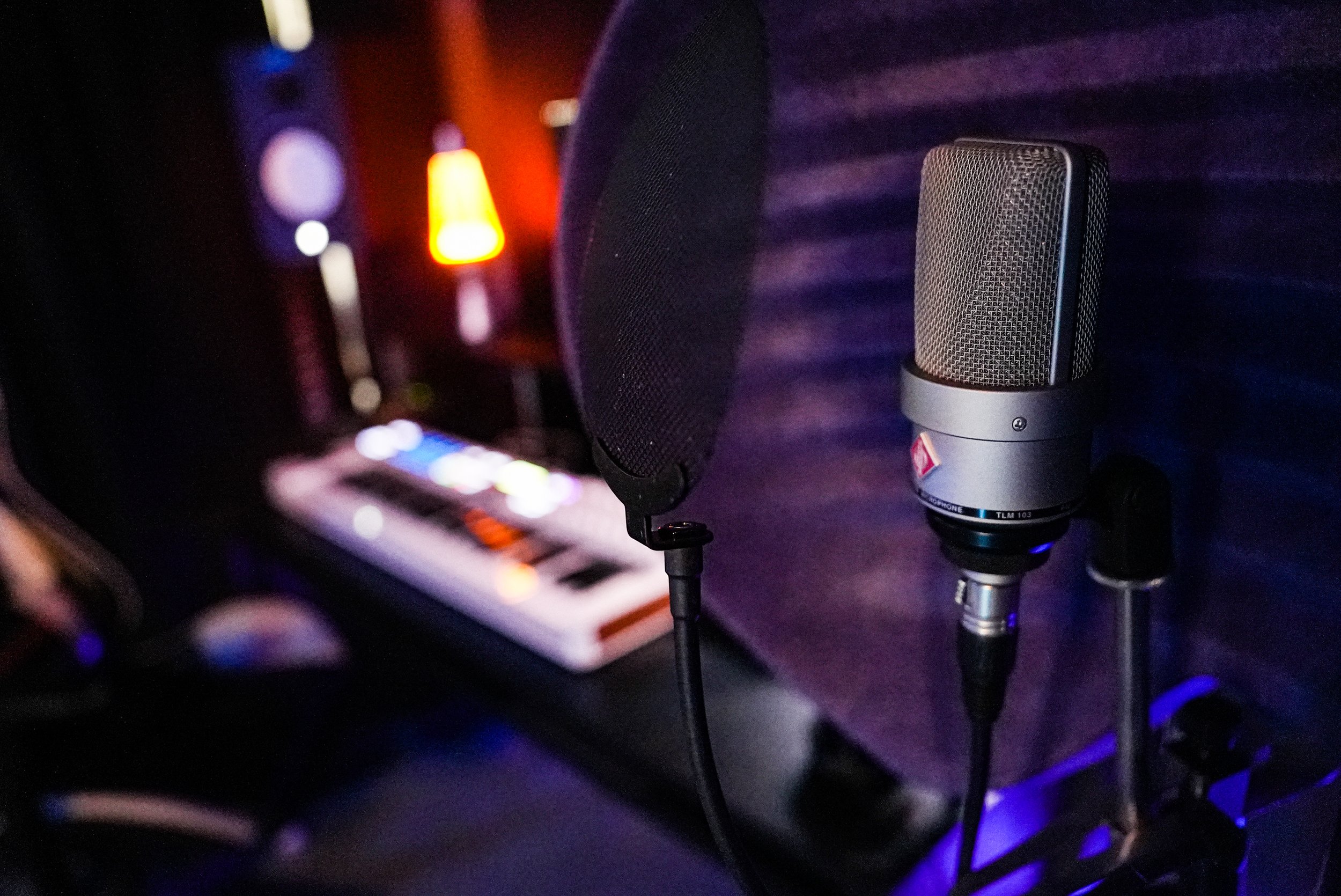 Studio microphone mounted on a stand, with a pop filter in front of it and a keyboard in the background, illuminated by warm and cool colored lights.