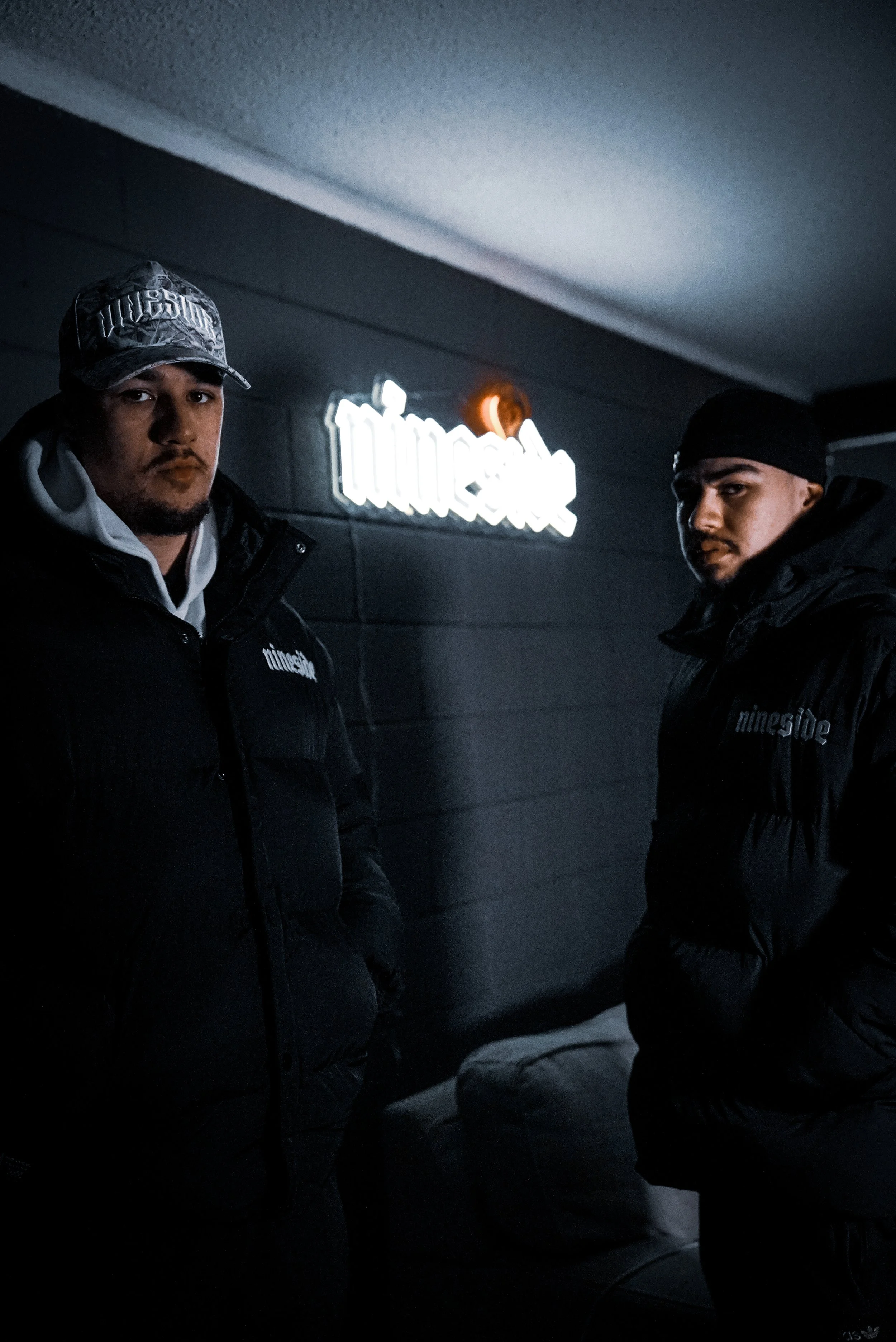 Two men standing in a dark room with a illuminated 'tuneside' sign on the black wall behind them, wearing black puffer jackets.