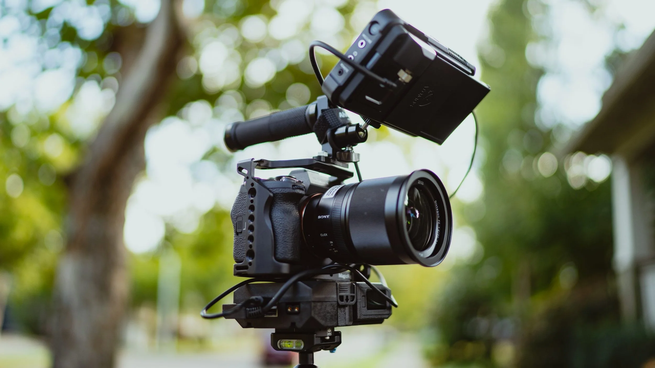 A professional video camera mounted on a stabilizer, equipped with a mounted external monitor, outdoors with blurred green trees in the background.