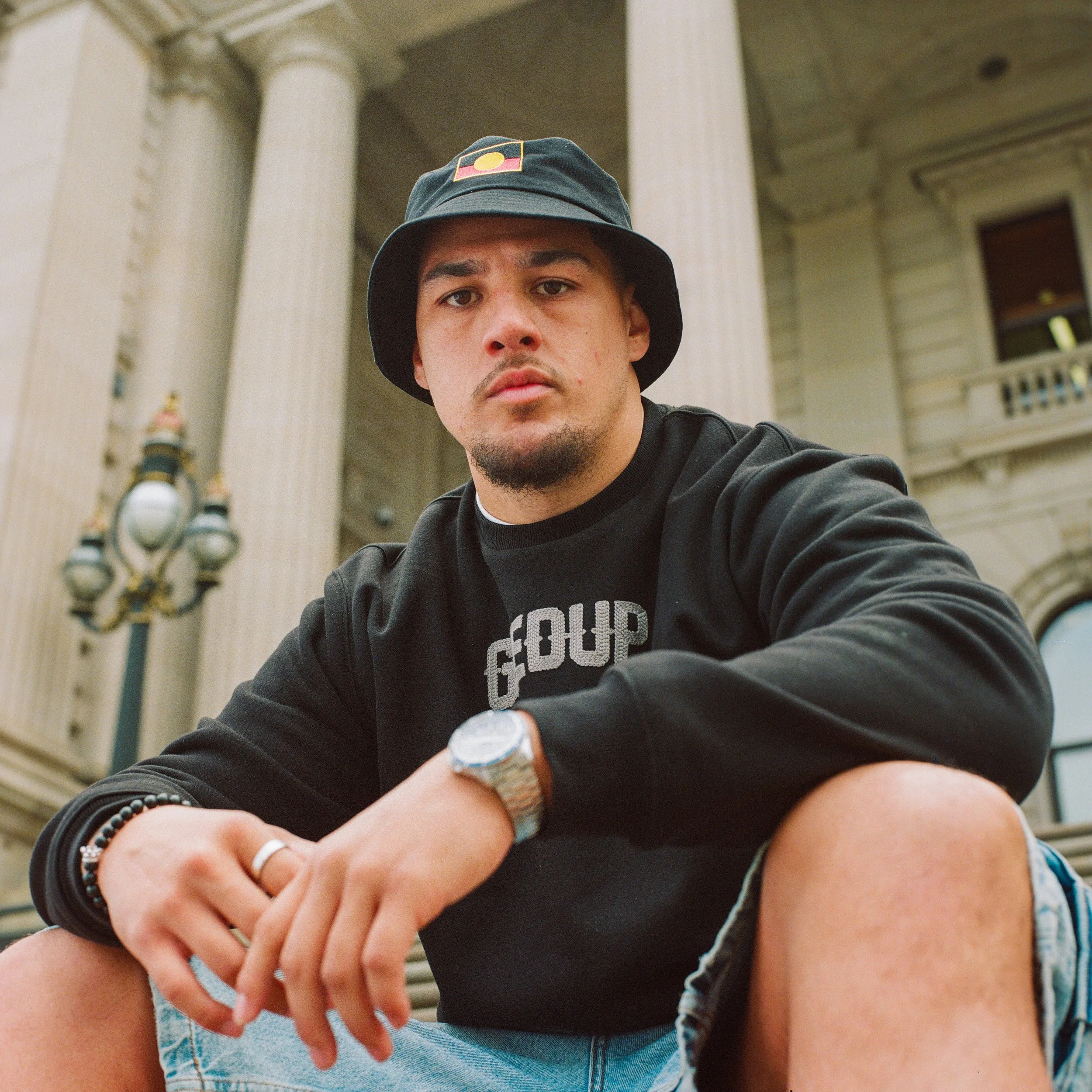 Young man sitting outdoors in front of a grand building with tall columns, wearing a bucket hat, black sweatshirt, and denim shorts, looking at the camera.