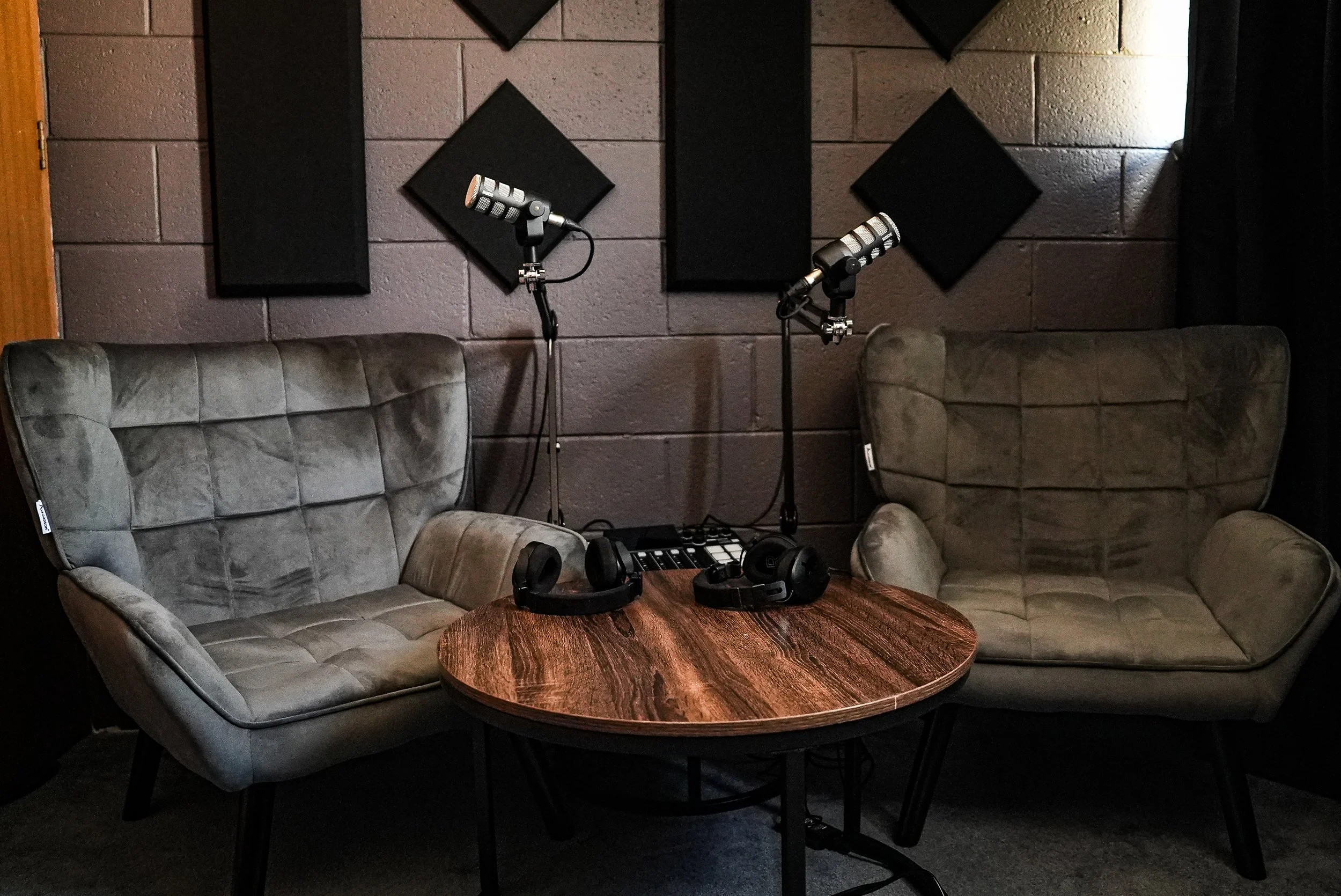 Recording studio setup with two gray upholstered armchairs, a round wooden table with headphones, microphones on stands, and soundproofing foam on the wall.